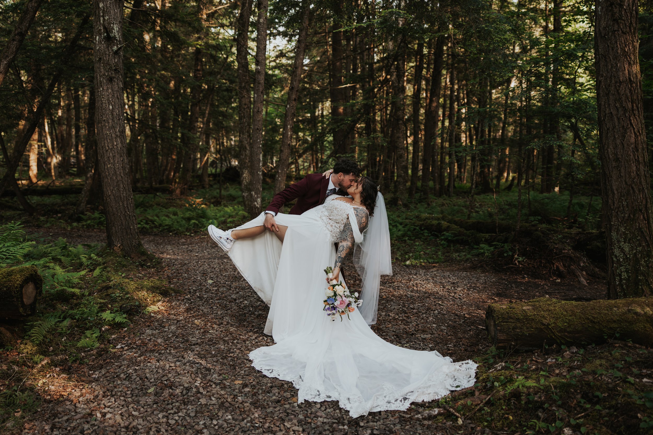 A newlywed couple sharing a kiss in a forest, the groom dressed in a maroon suit and the bride in a white wedding dress, holding a bouquet of flowers.