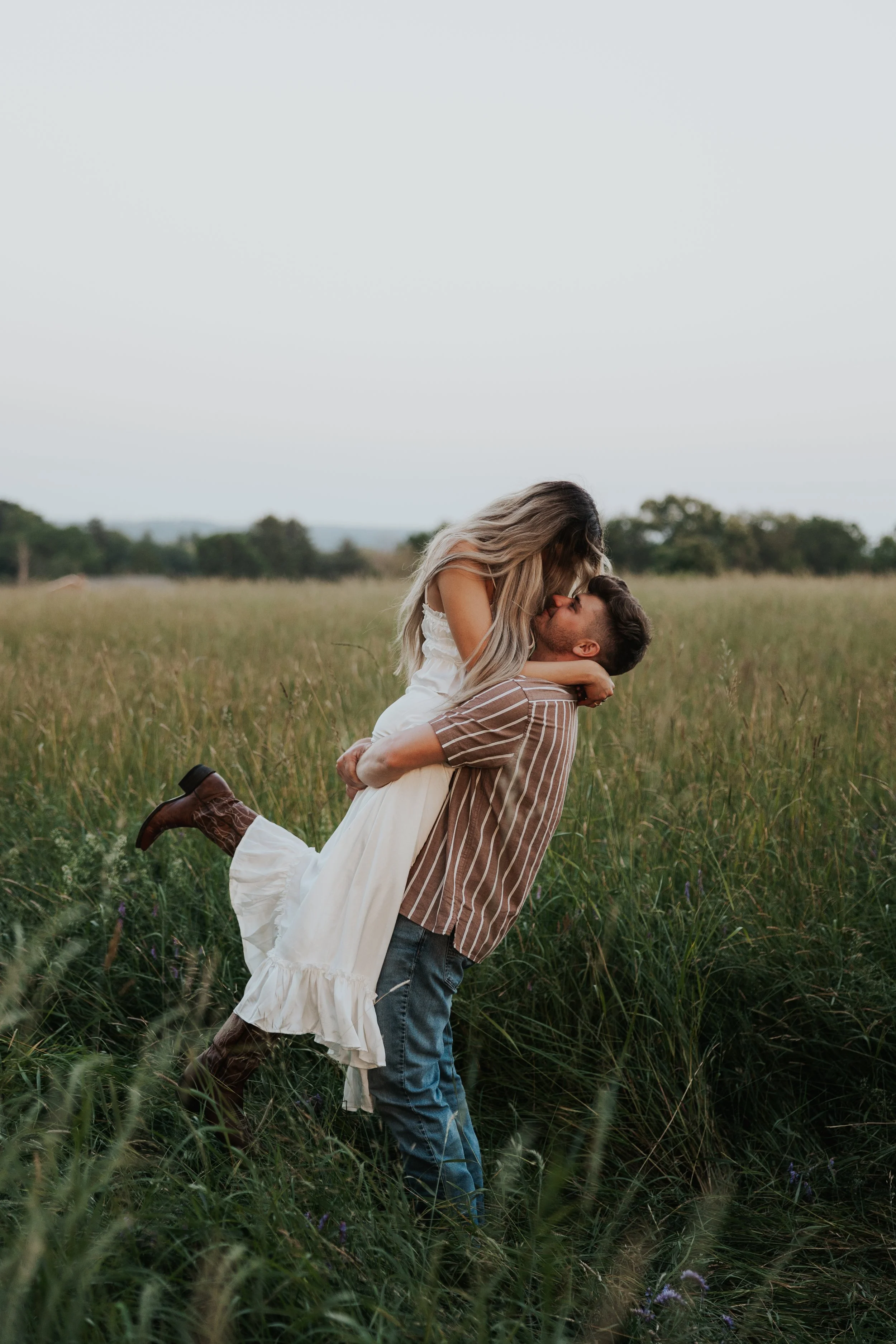 A couple outdoors in a field, with the man lifting the woman in his arms, both kissing.