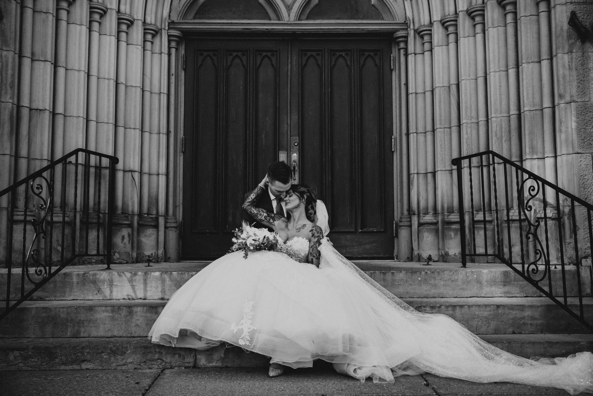 A bride and groom sitting on stone stairs in front of a large wooden door, embracing affectionately, with the bride wearing a wedding gown and holding a bouquet, captured in black and white.