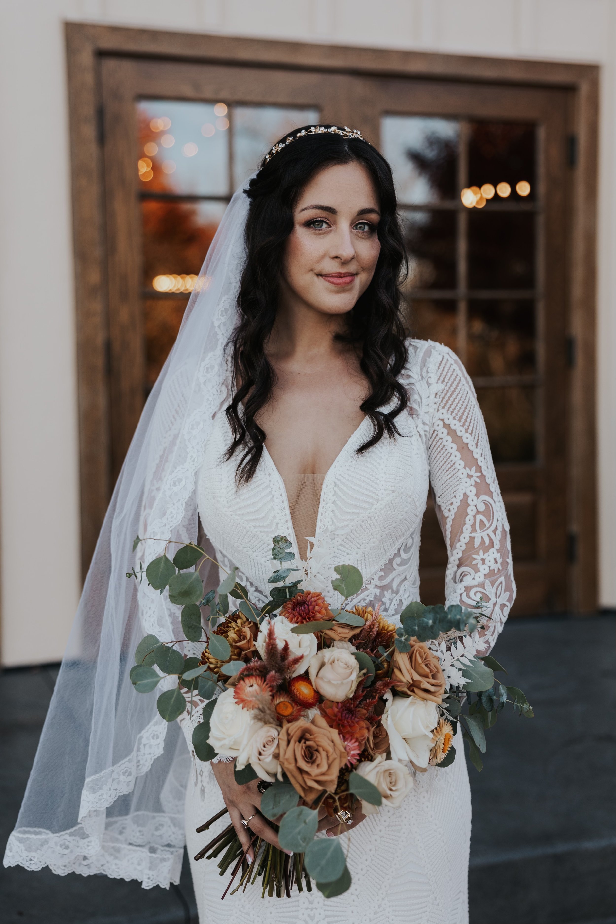 Bride with dark wavy hair in a white lace wedding gown holding a bouquet of white and tan roses with green leaves, standing indoors in front of a wooden door with glass panels and blurred outdoor lights in the background.