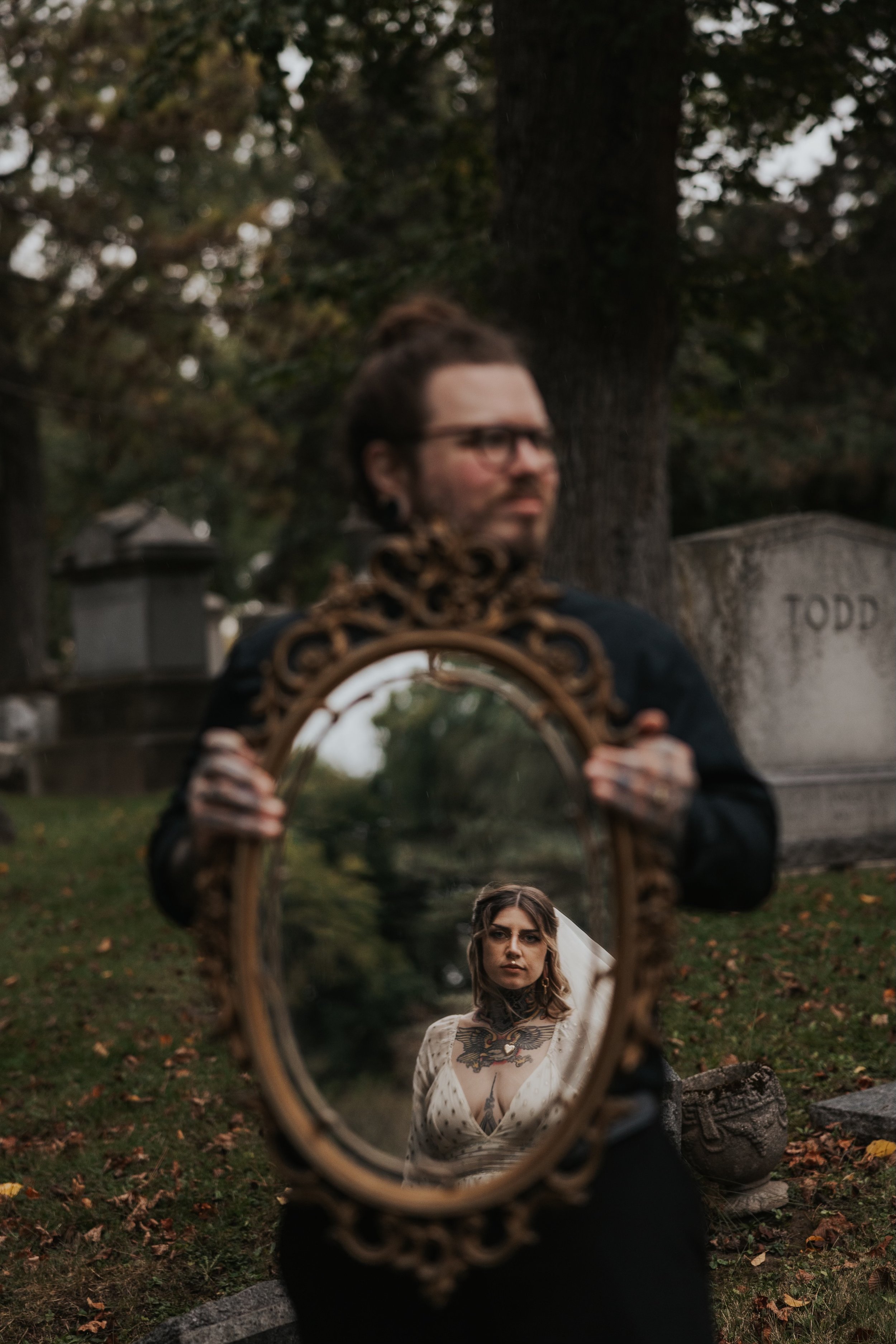 A person holding an ornate mirror reflecting a woman with tattoos on her chest and shoulders, standing outdoors in a cemetery with gravestones and trees in the background.