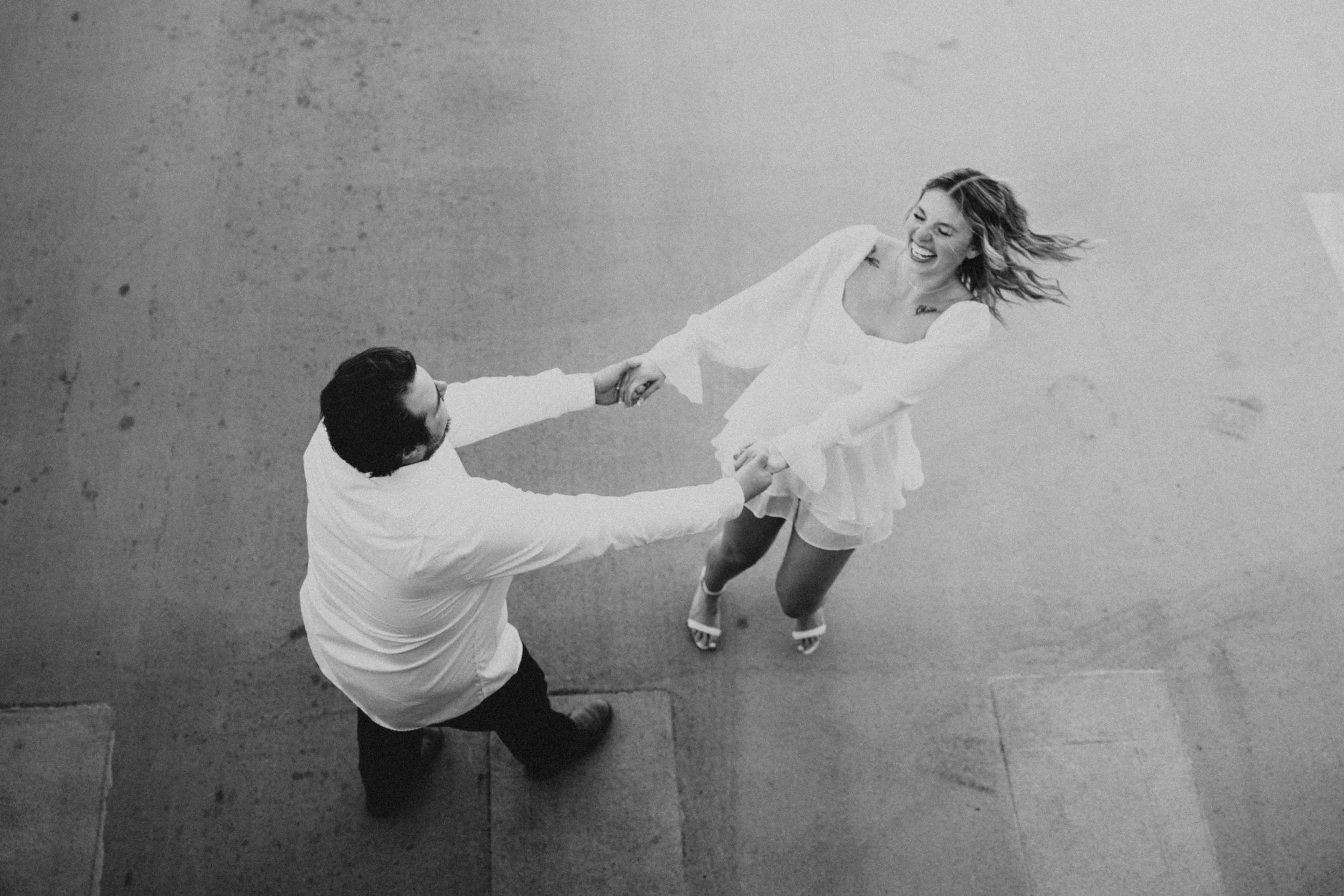 A black-and-white photo of a happy couple holding hands and dancing, viewed from above. The woman is smiling and wearing a light-colored dress, and the man is wearing a white shirt and dark pants.