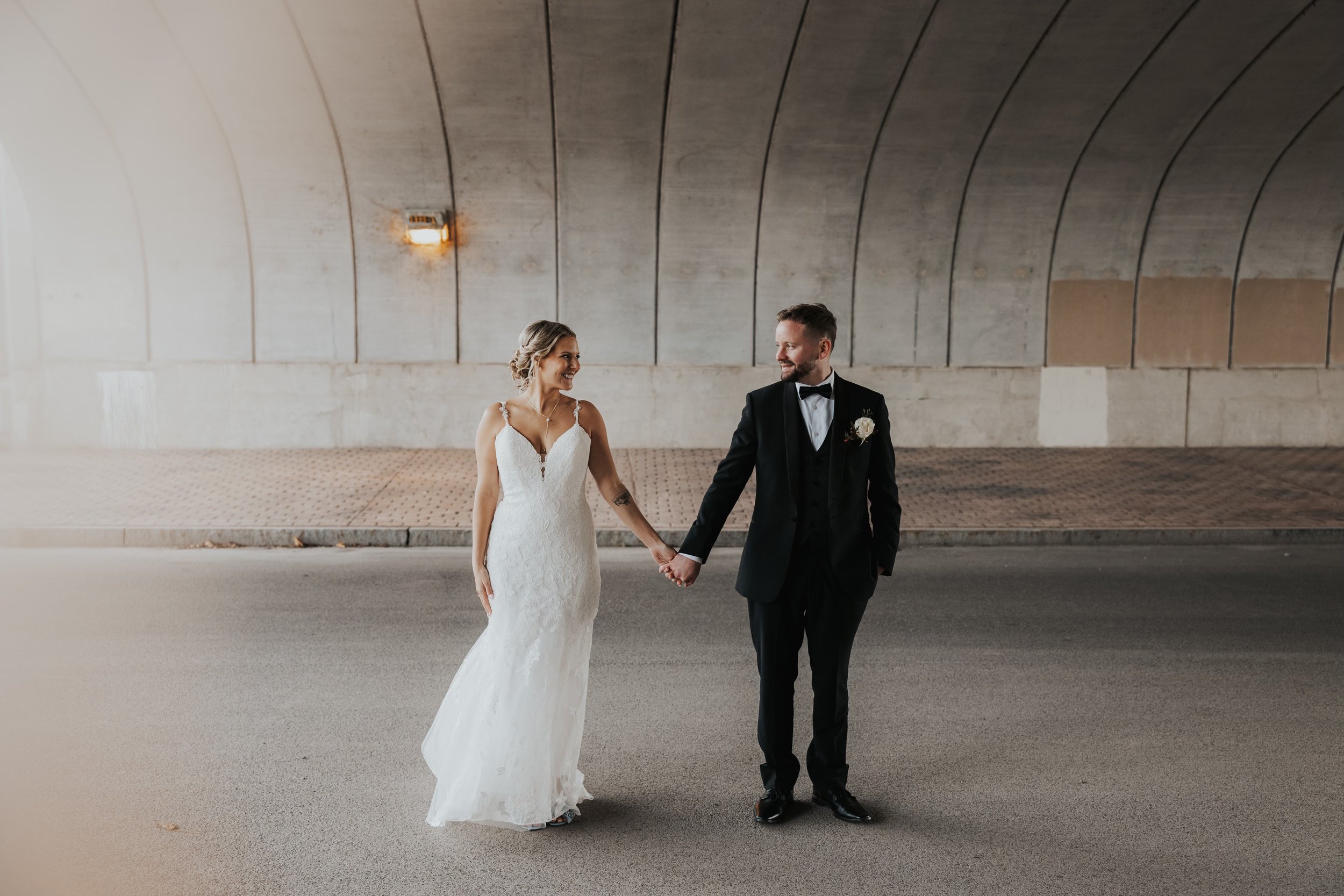 A bride and groom holding hands and smiling at each other under a concrete bridge