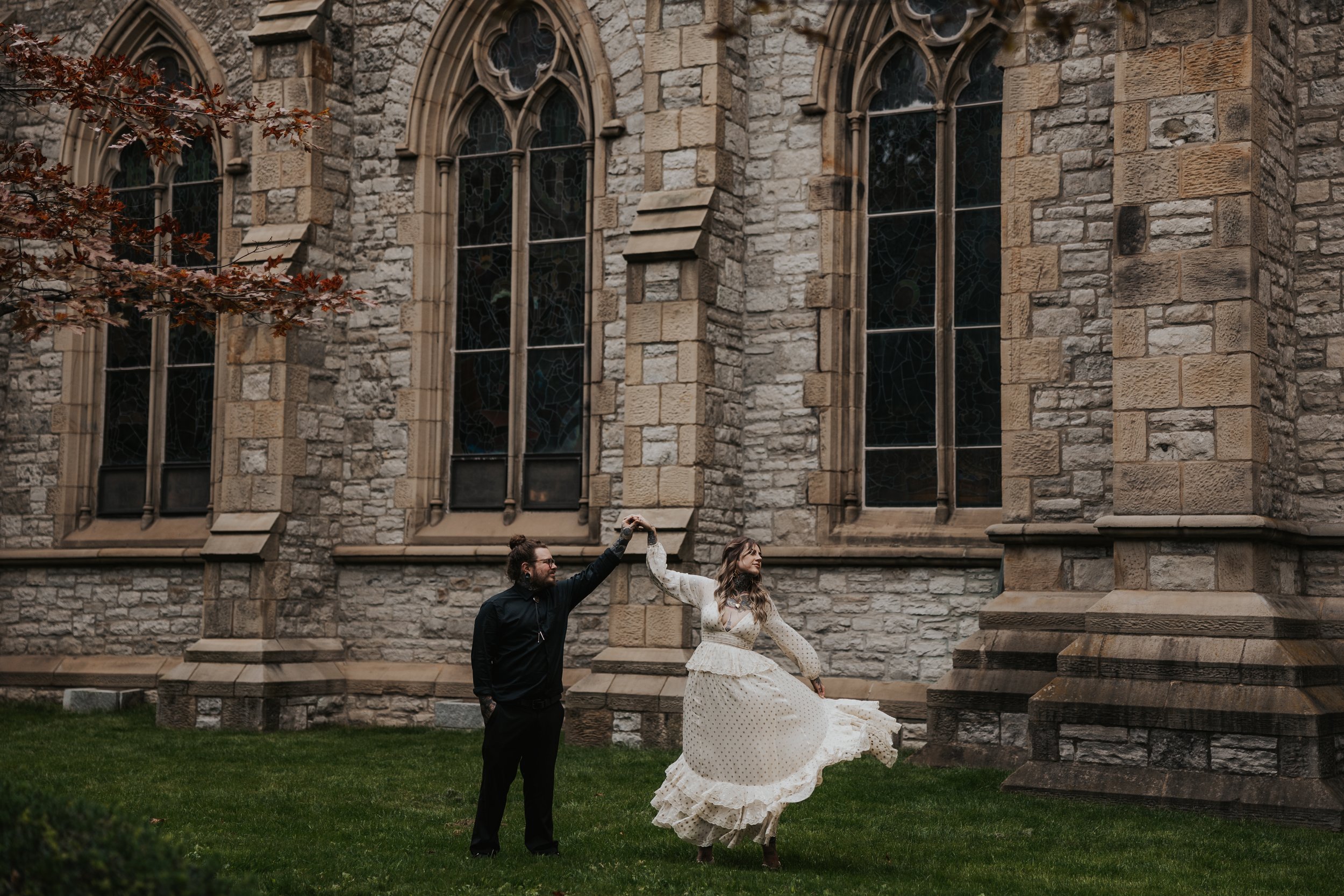 A man and woman dancing outside a stone church with large stained glass windows; the woman is twirling in a vintage-style white dress, and the man is holding her hand.