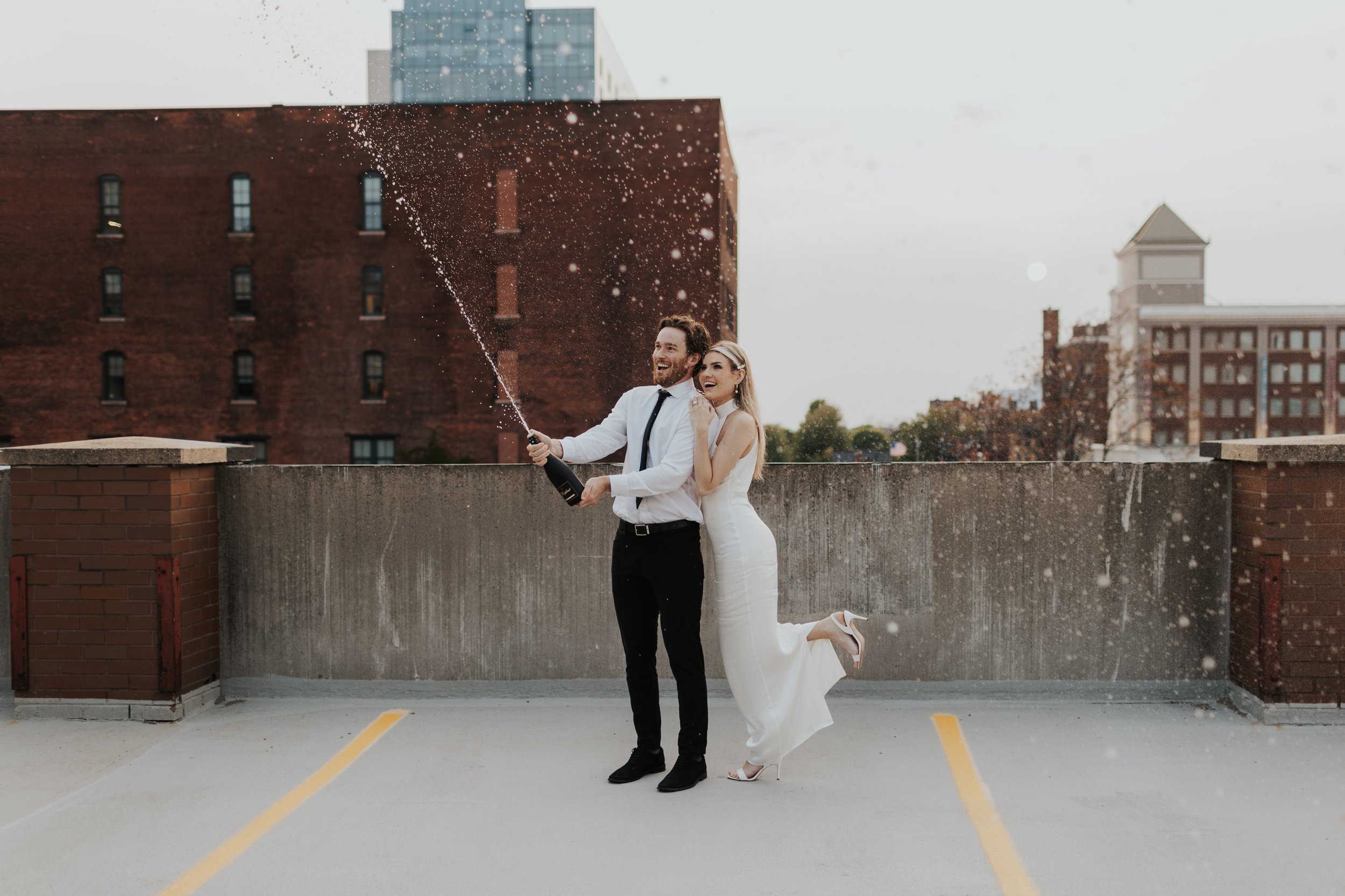 A bride and groom celebrating on a rooftop parking lot, with the groom opening a champagne bottle and the bride joyfully jumping, as champagne sprays into the air, with city buildings in the background.