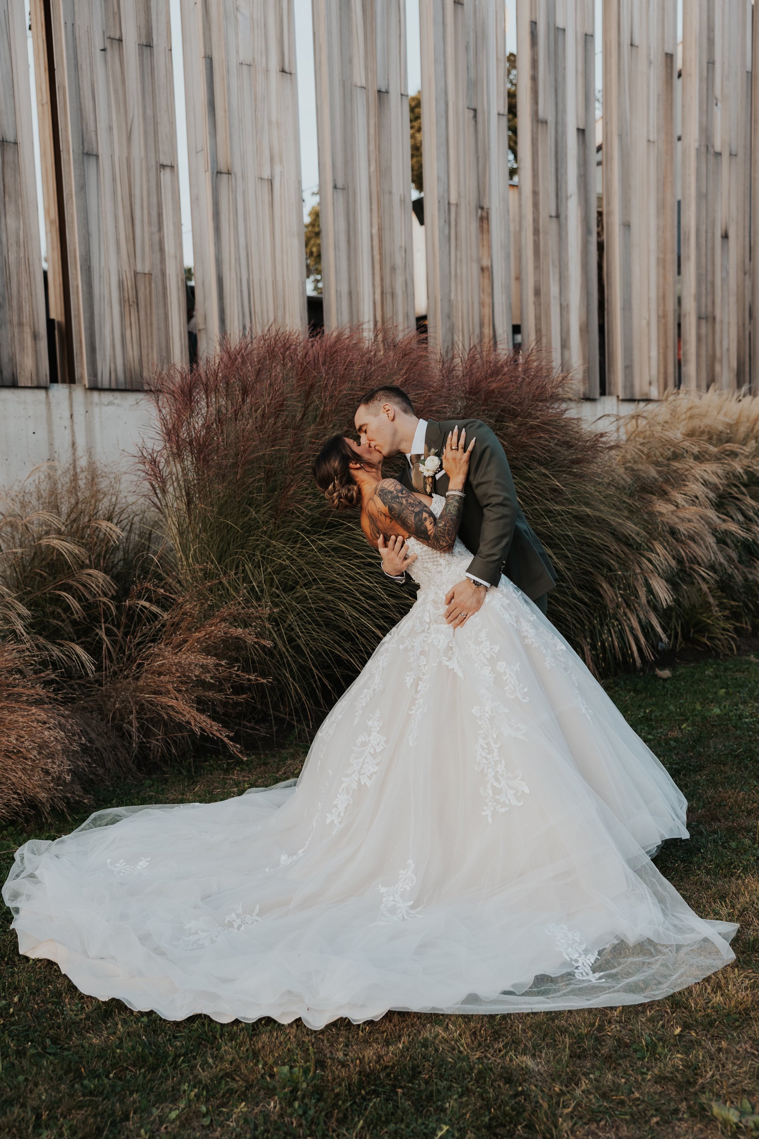 A newlywed couple shares a romantic kiss outdoors, the bride in a white lace wedding gown and the groom in a dark suit, standing on grass in front of ornamental grasses and a wooden structure.