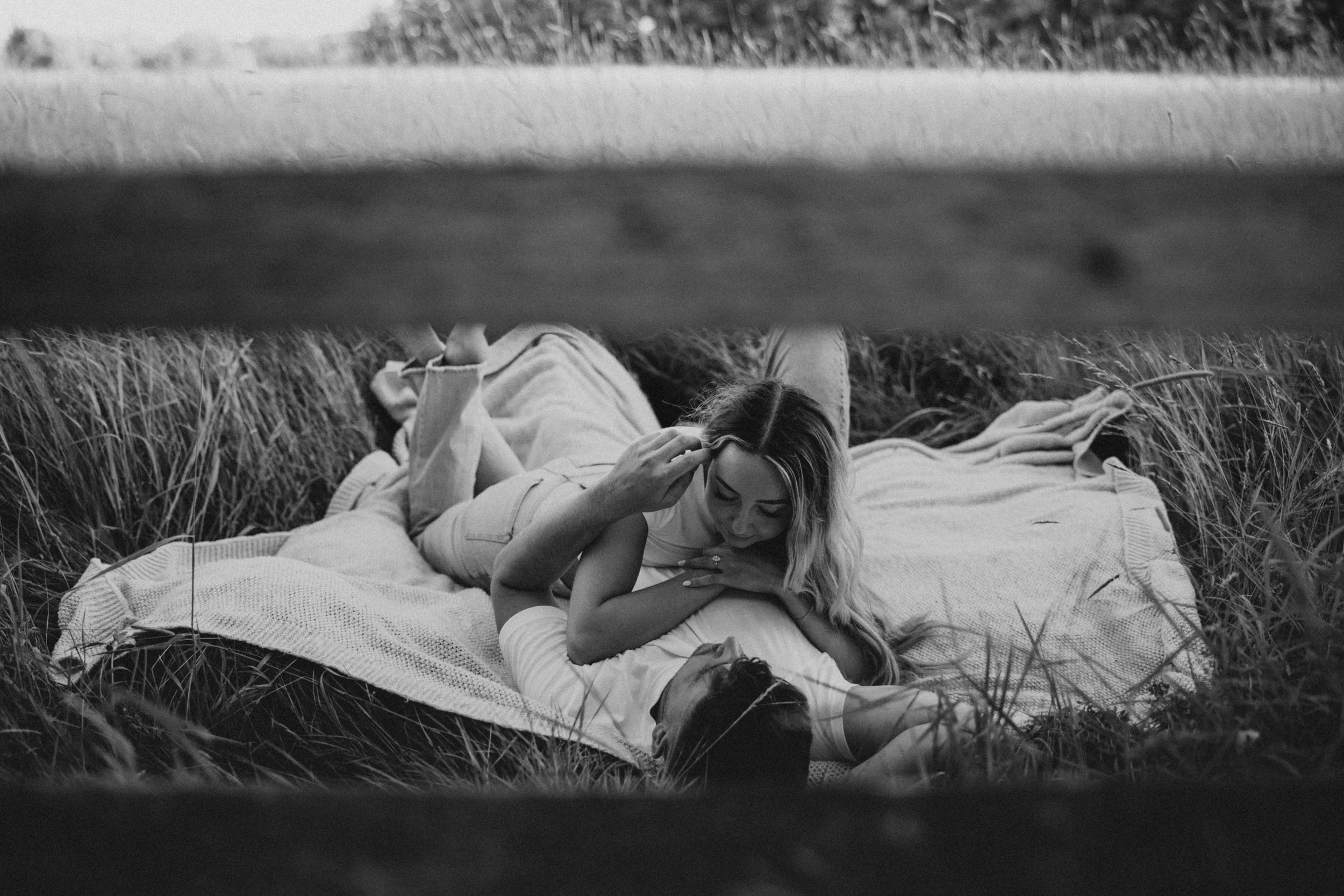 A woman and man lying on a blanket in tall grass, engaging in an intimate moment outdoors, viewed from beneath a wooden structure.