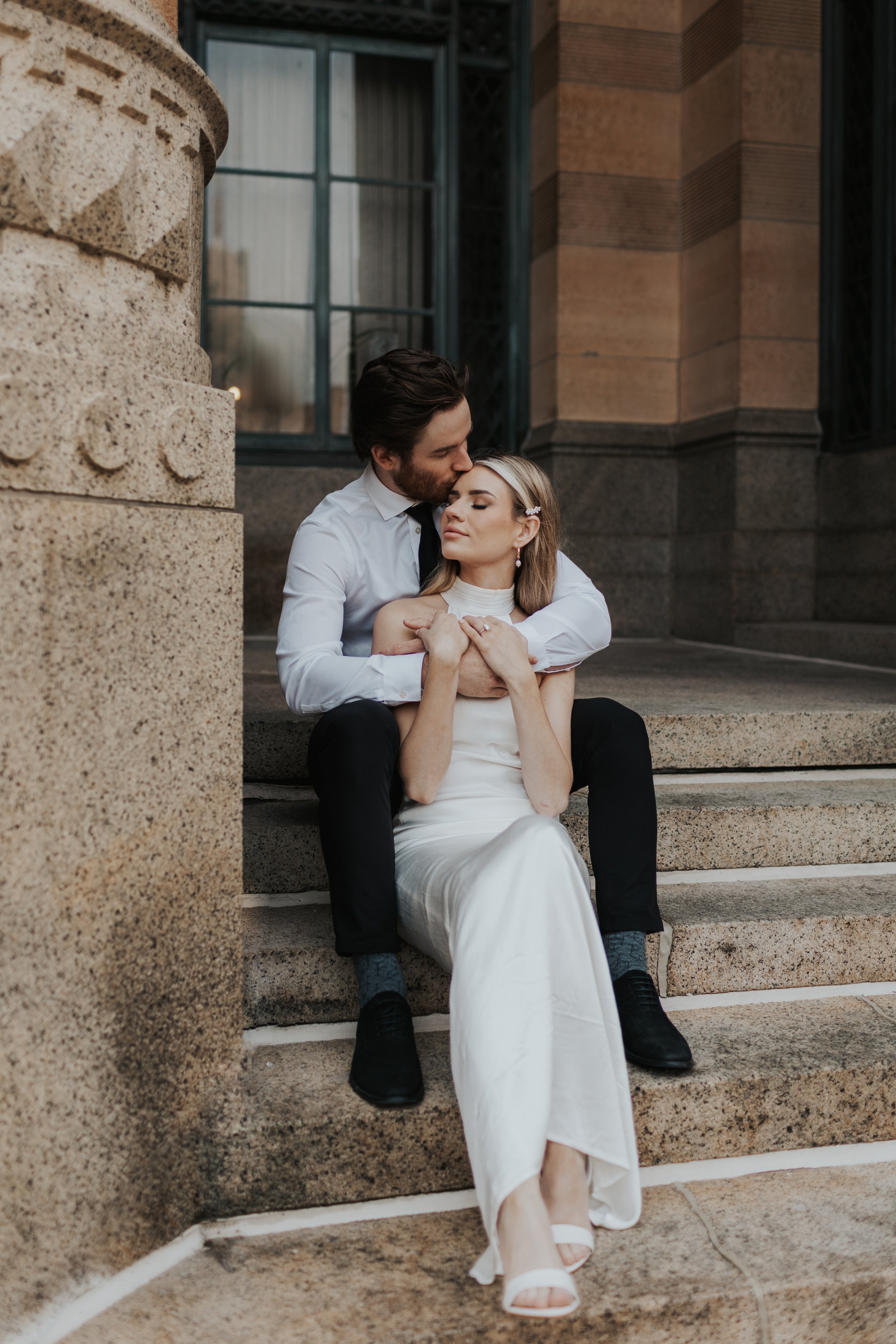A couple sitting on steps outside a building, with the man embracing the woman from behind, kissing her forehead. The woman is wearing a white dress and the man a white shirt and black pants.