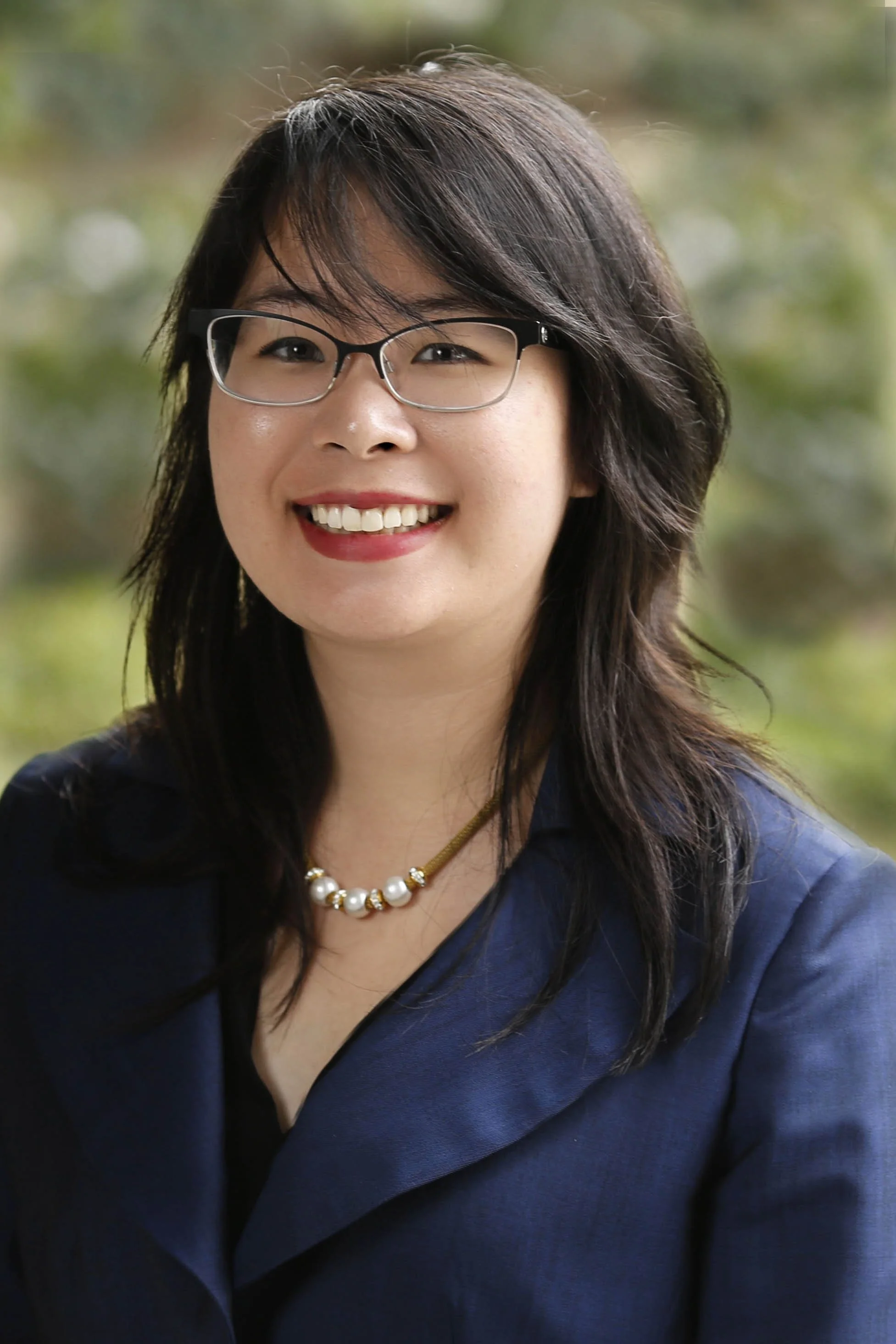 A smiling woman with glasses and dark hair, wearing a navy blazer and pearl necklace, outdoors with a blurred green background.