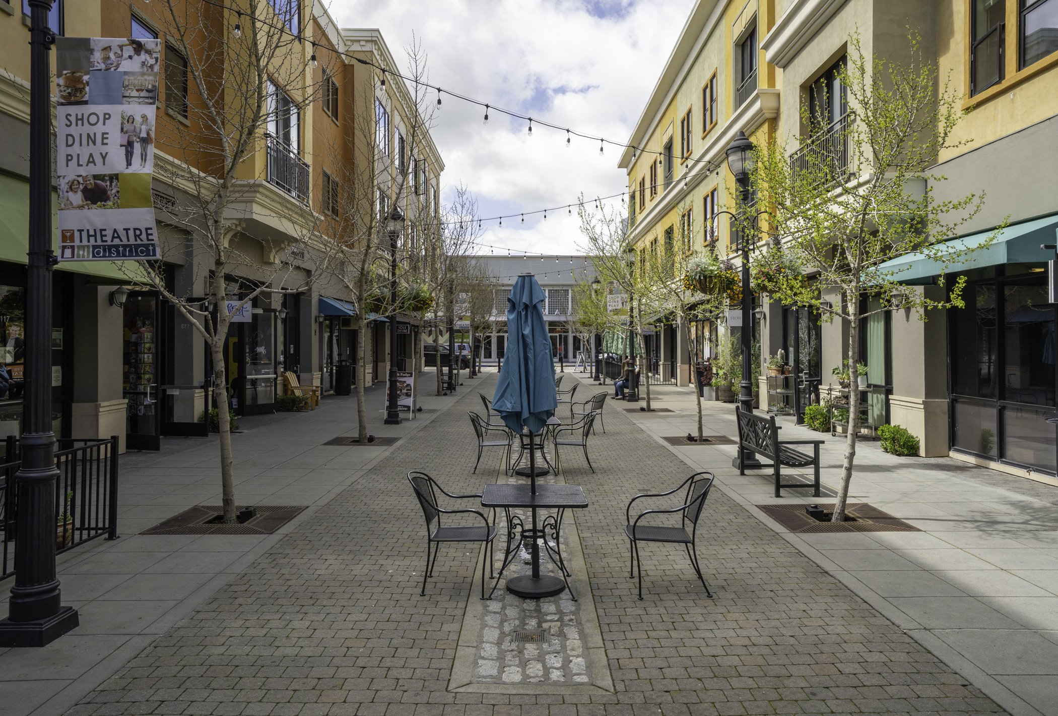 Empty outdoor shopping area with dining tables, chairs, a blue umbrella, string lights, trees, and shops on both sides.