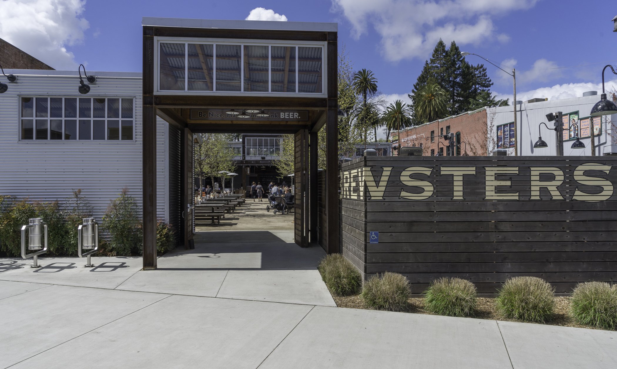 Outdoor seating area with wooden tables and benches, surrounded by modern industrial buildings and signage. Trees and a clear blue sky in the background.