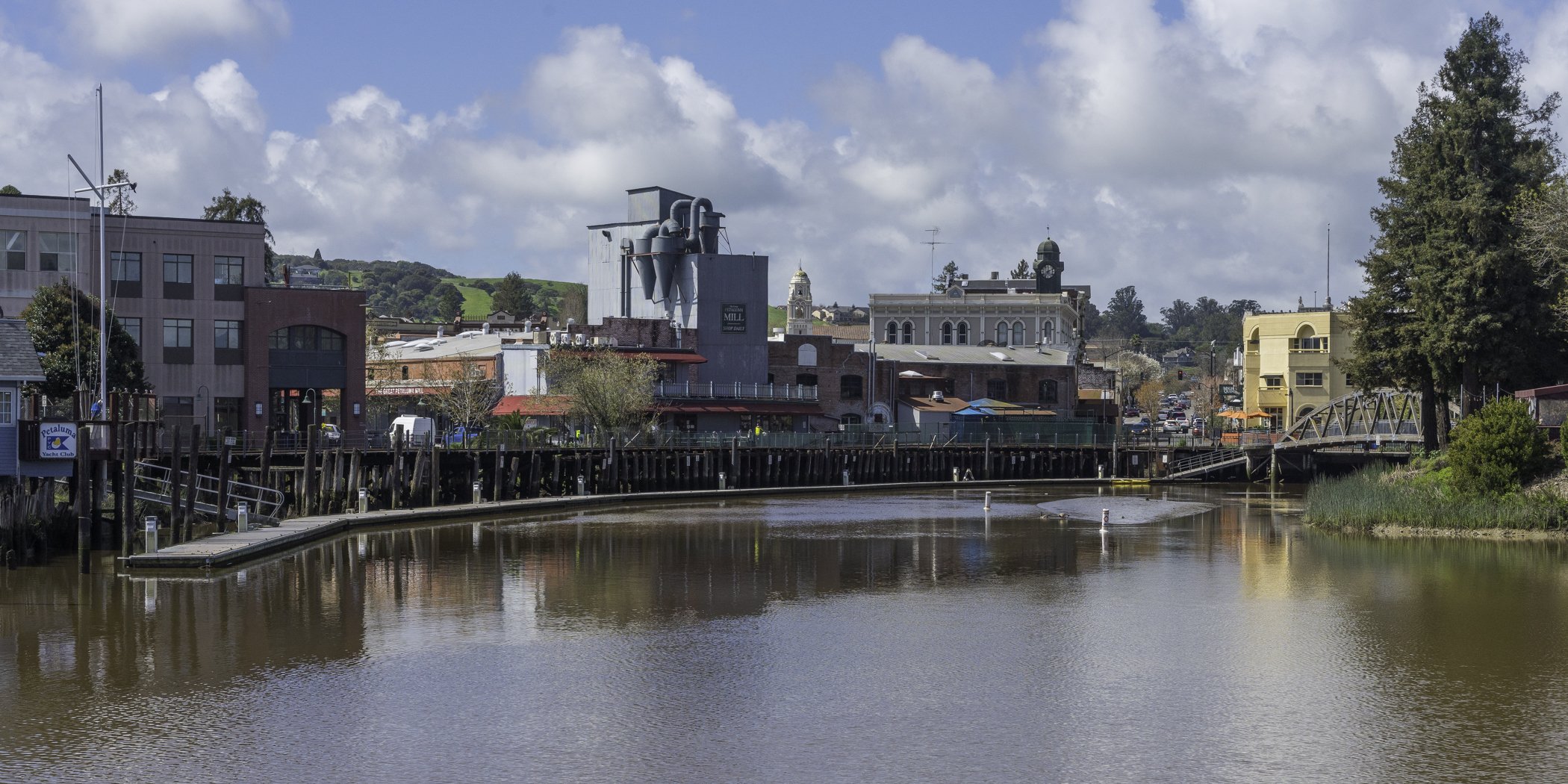 Riverside town with historic buildings and a bridge in Petaluma, California, reflecting in the water under a partly cloudy sky.