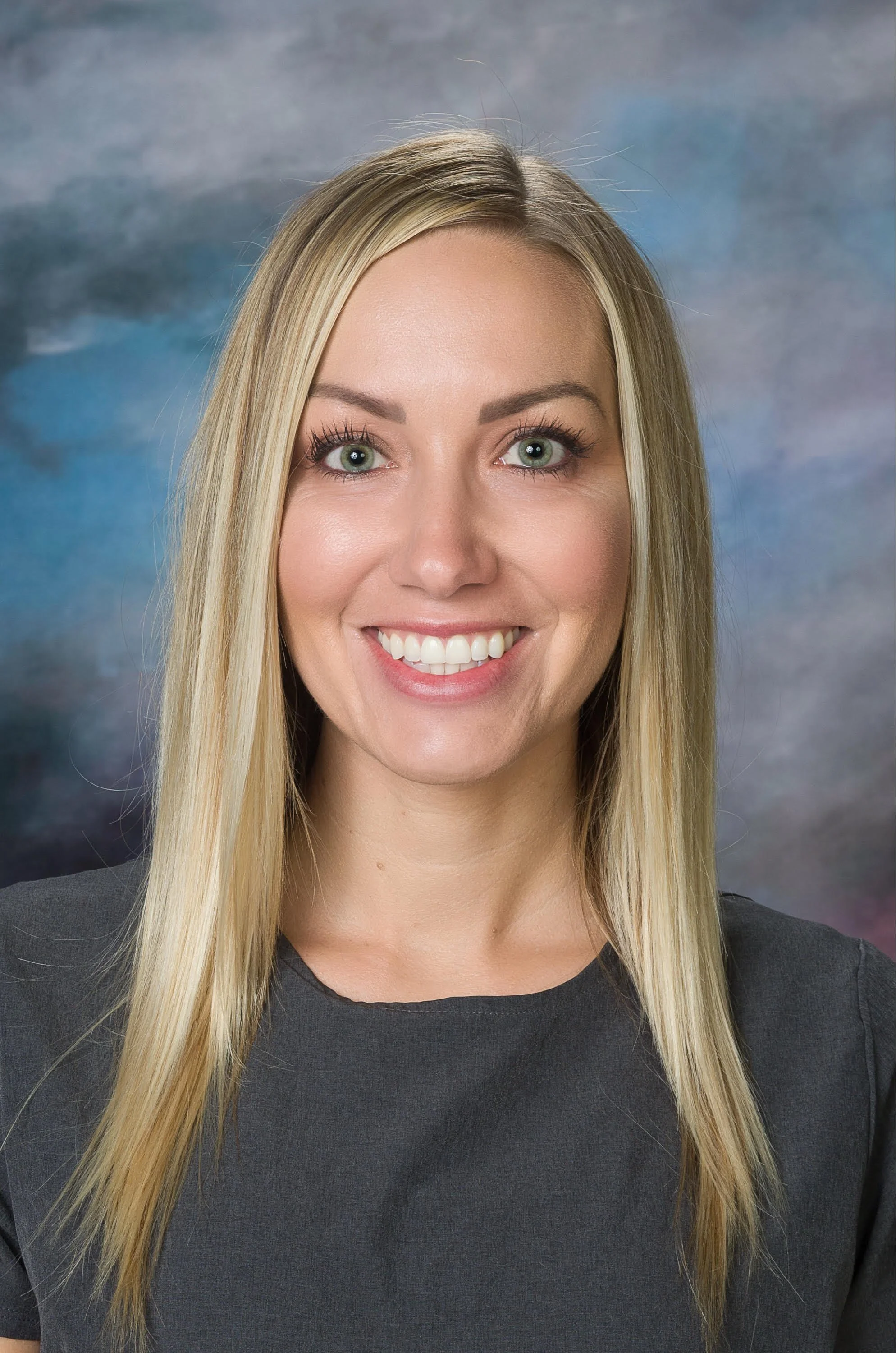 Headshot of a woman with long blonde hair, green eyes, and a black top, smiling, against a cloudy sky background.