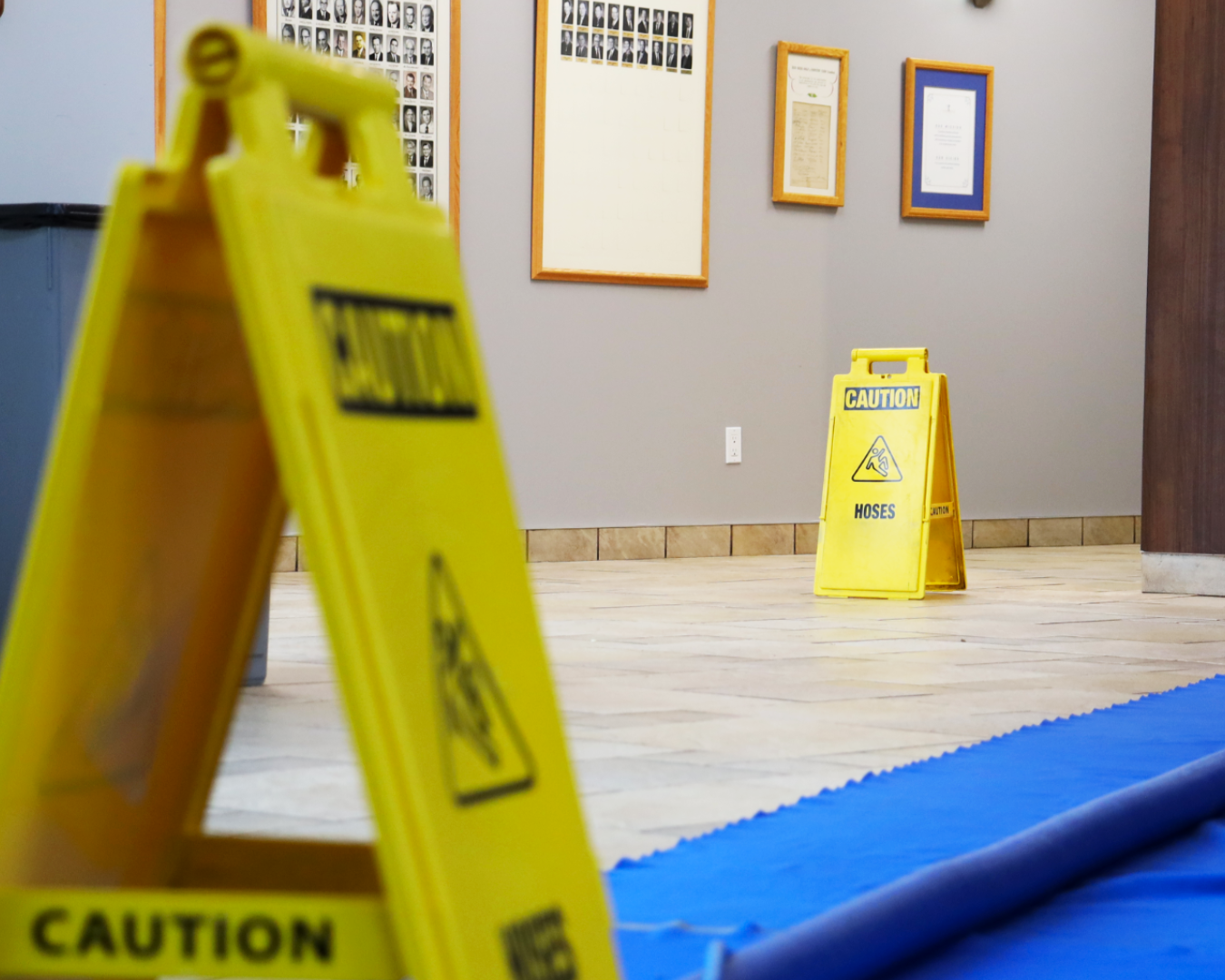 Yellow caution signs indicating wet floor and hose warnings in an indoor space with framed pictures and certificates on the wall, and blue flooring mat.