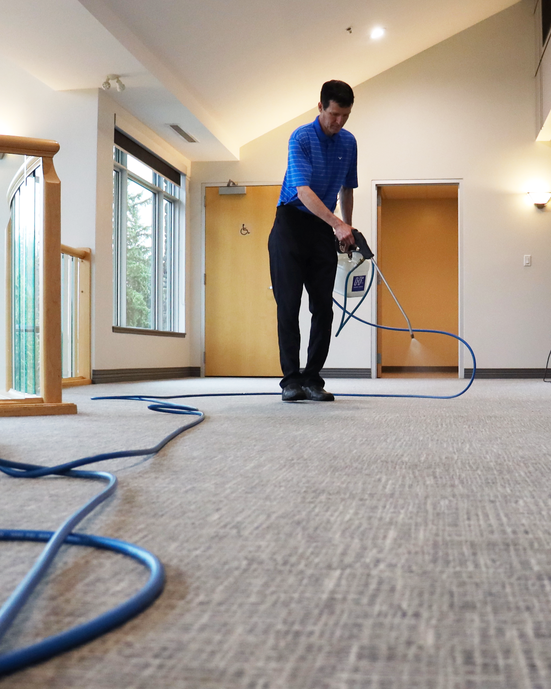 A man wearing a blue polo shirt and black pants is using a steam cleaner to clean a beige carpet in a well-lit indoor space with large windows and wooden accents.