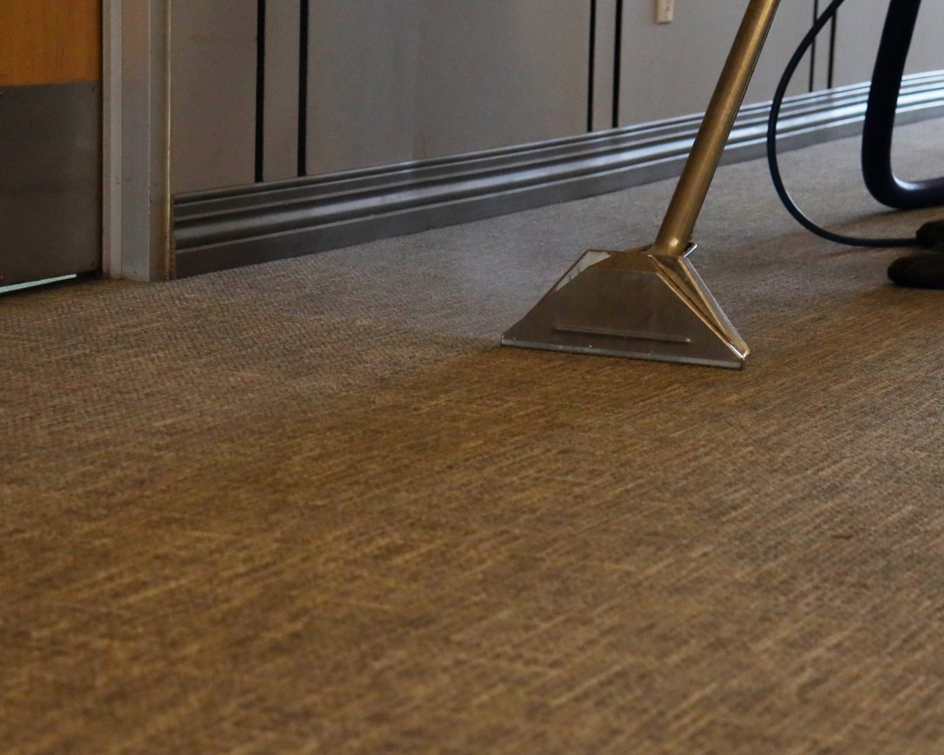 Close-up of a door bottom sweep with an angled metal piece on a brown carpeted floor near a metallic baseboard and door frame.
