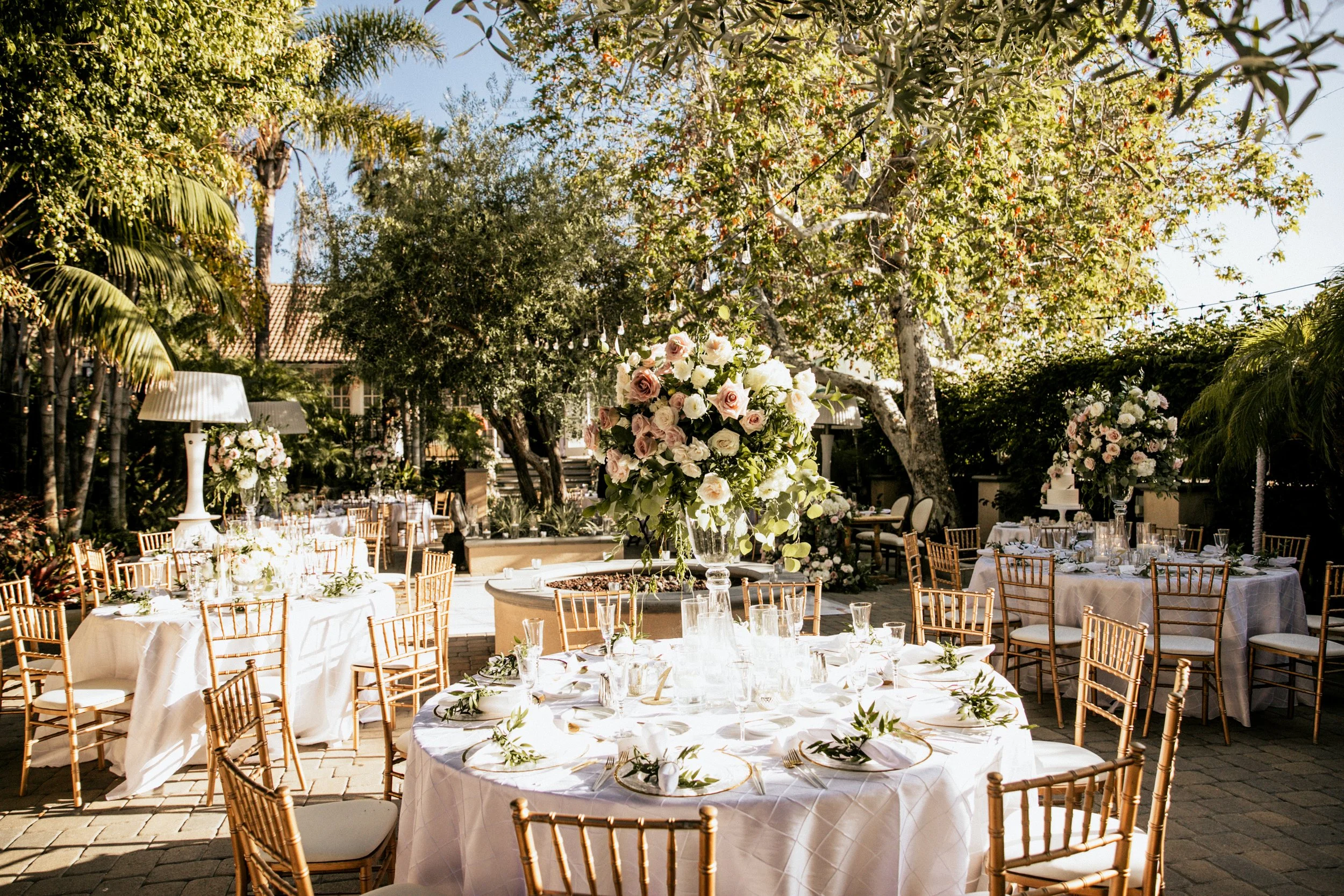 Elegant outdoor dining setup at a wedding with a dark wood table, clear chairs, floral arrangements, candles, and a chandelier under a white canopy with draped fabric.