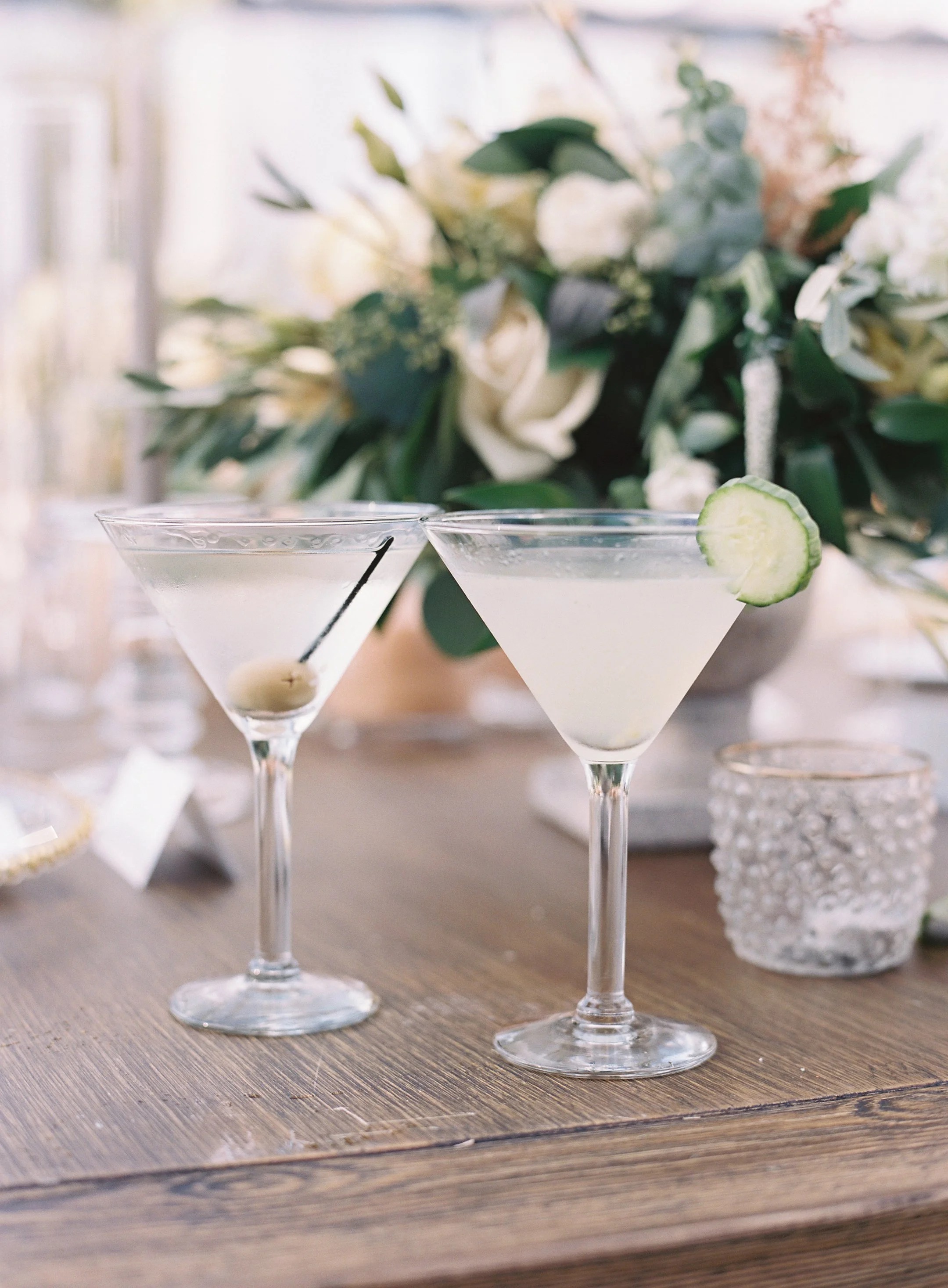 Elegant outdoor dining setup at a wedding with a dark wood table, clear chairs, floral arrangements, candles, and a chandelier under a white canopy with draped fabric.