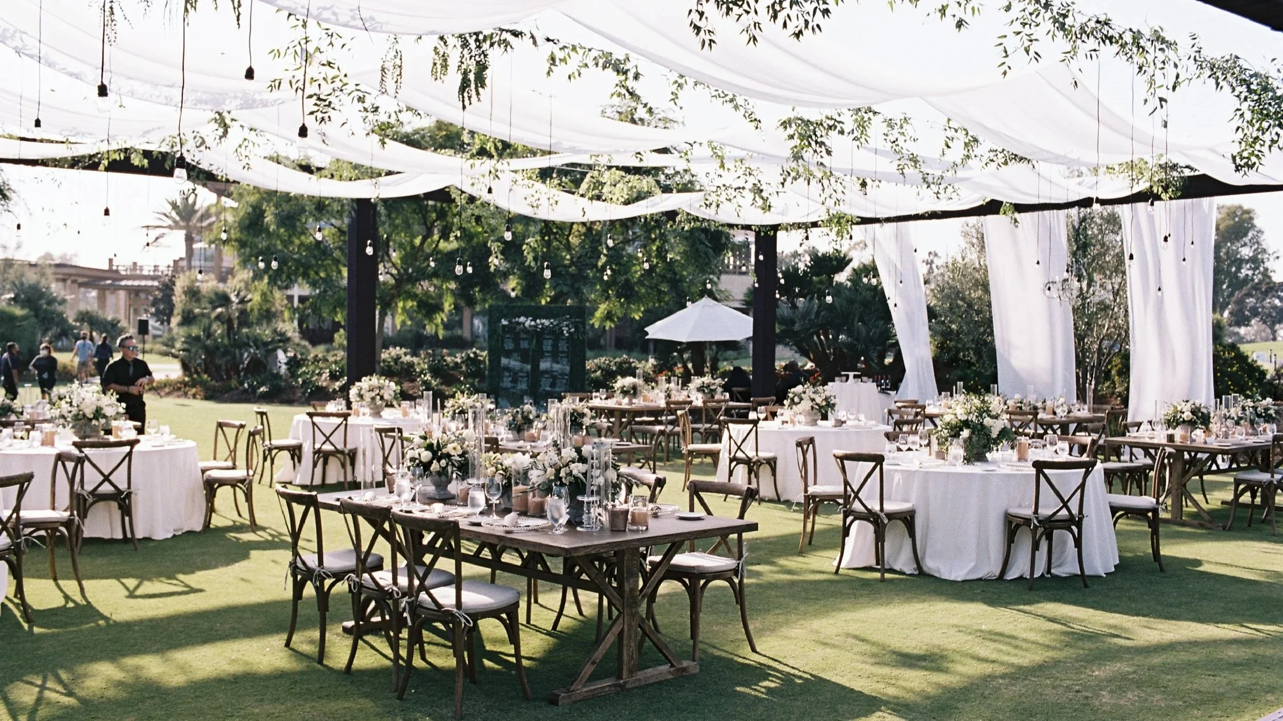 Outdoor garden setting with round and rectangular tables decorated with floral arrangements, surrounded by wooden chairs and lush tropical plants, including palm trees and large green leaves.