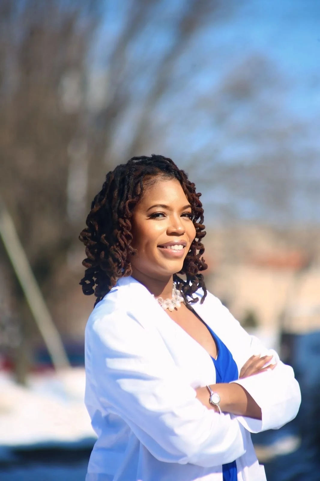 A woman with curly hair wearing a white blazer and blue top, standing outdoors with her arms crossed, smiling at the camera.