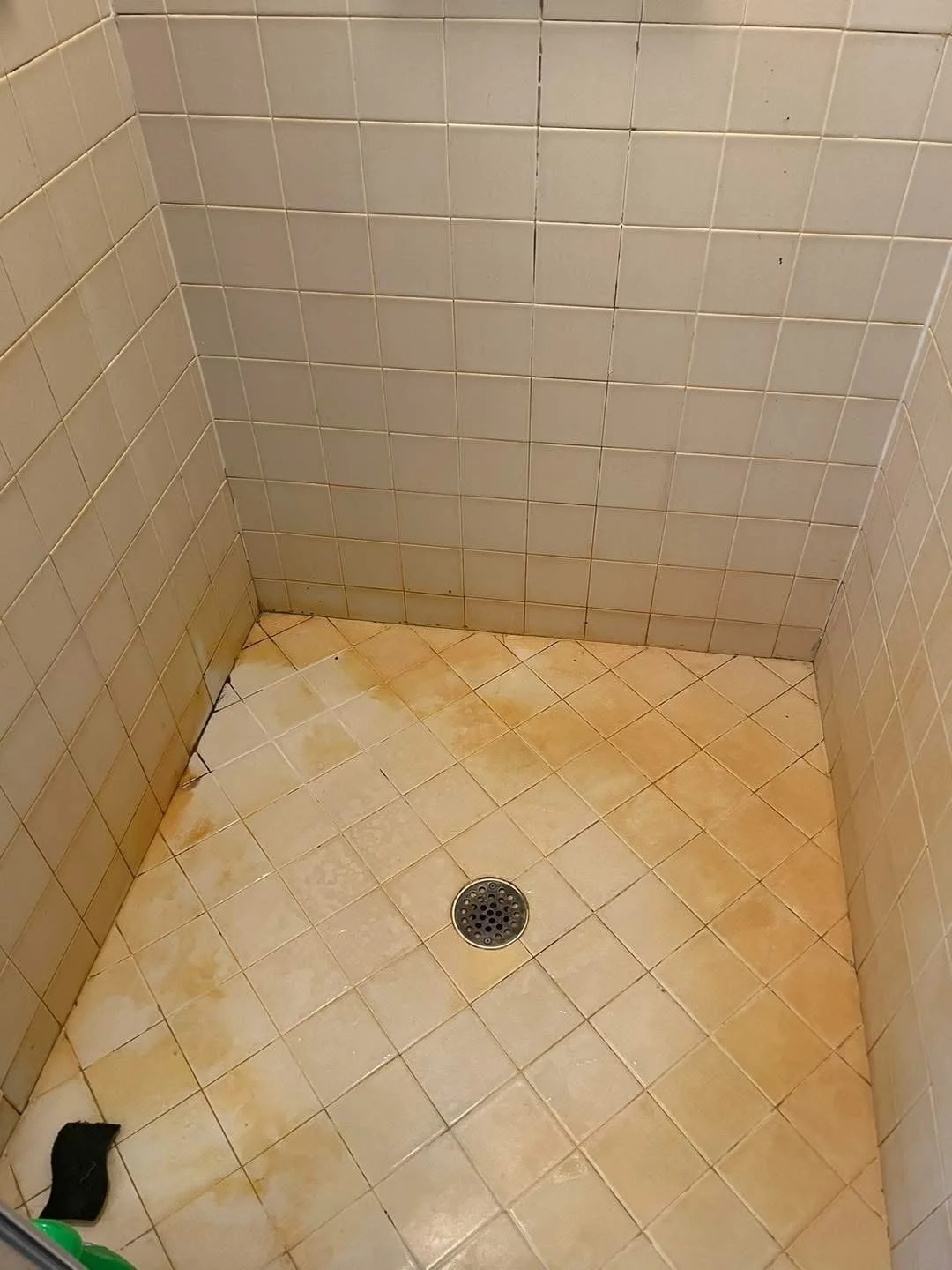 Dirty bathroom shower floor with yellowish stains, a drain in the center, tiled walls, and a black scrub sponge in the lower left corner.