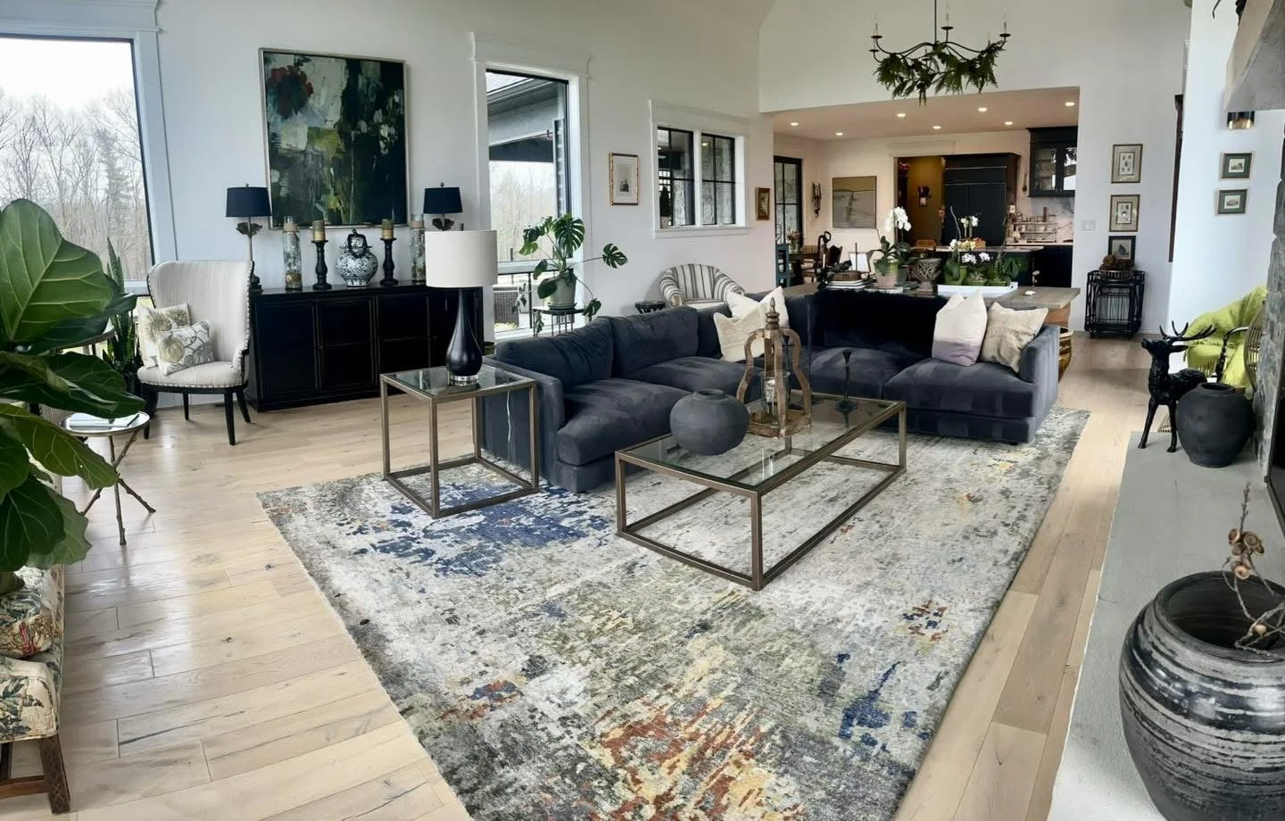 Modern living room with a large dark sectional sofa, glass coffee table, and various decorative items, large windows, and light wood flooring.