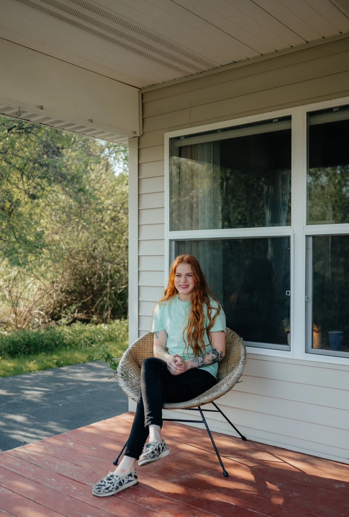 A red-haired woman with tattoos sitting on a wicker chair on an outdoor porch with a house and green trees in the background.