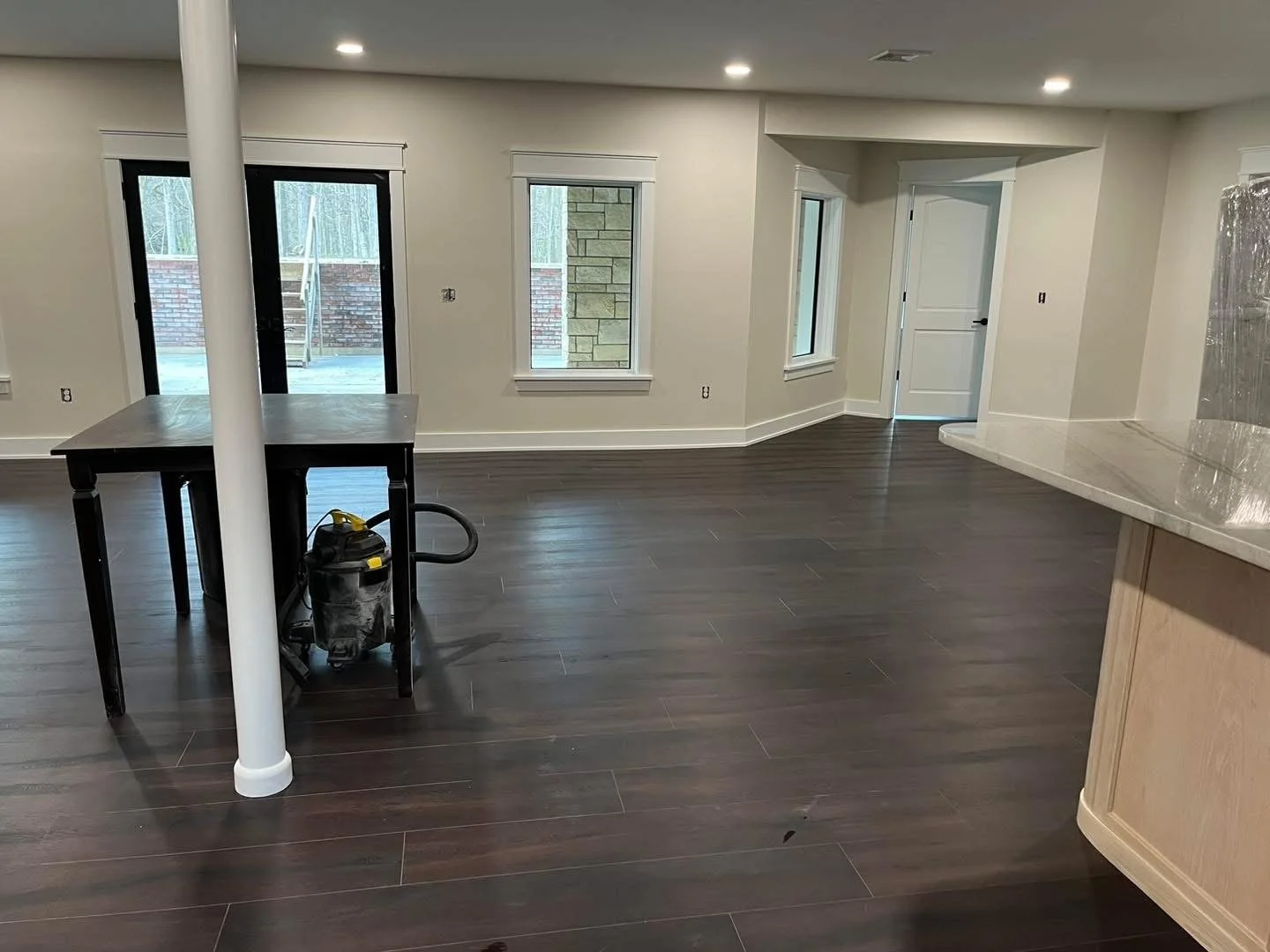 Empty modern living space with dark wood flooring, white walls, two windows, a door, and a small kitchen countertop on the right. There is also a table with a vacuum cleaner underneath, a white support pole, and recessed lighting.