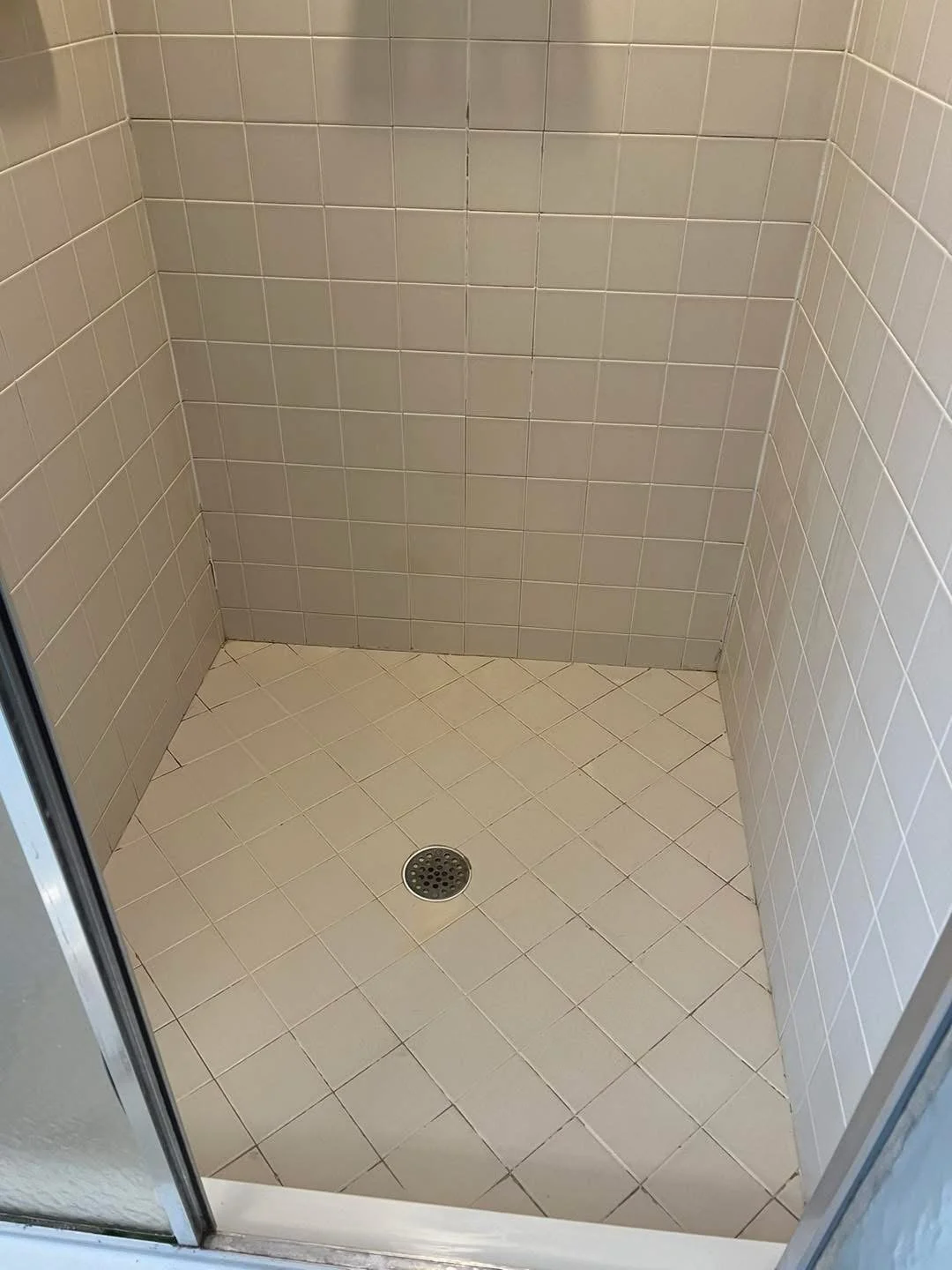 Empty shower stall with beige tiled walls and floor, metal drain in the center, and a glass door on the left side.