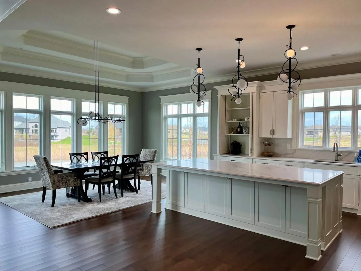 Open-concept kitchen and dining area with large windows, white cabinetry, a marble island, dark wood flooring, and modern pendant lighting.