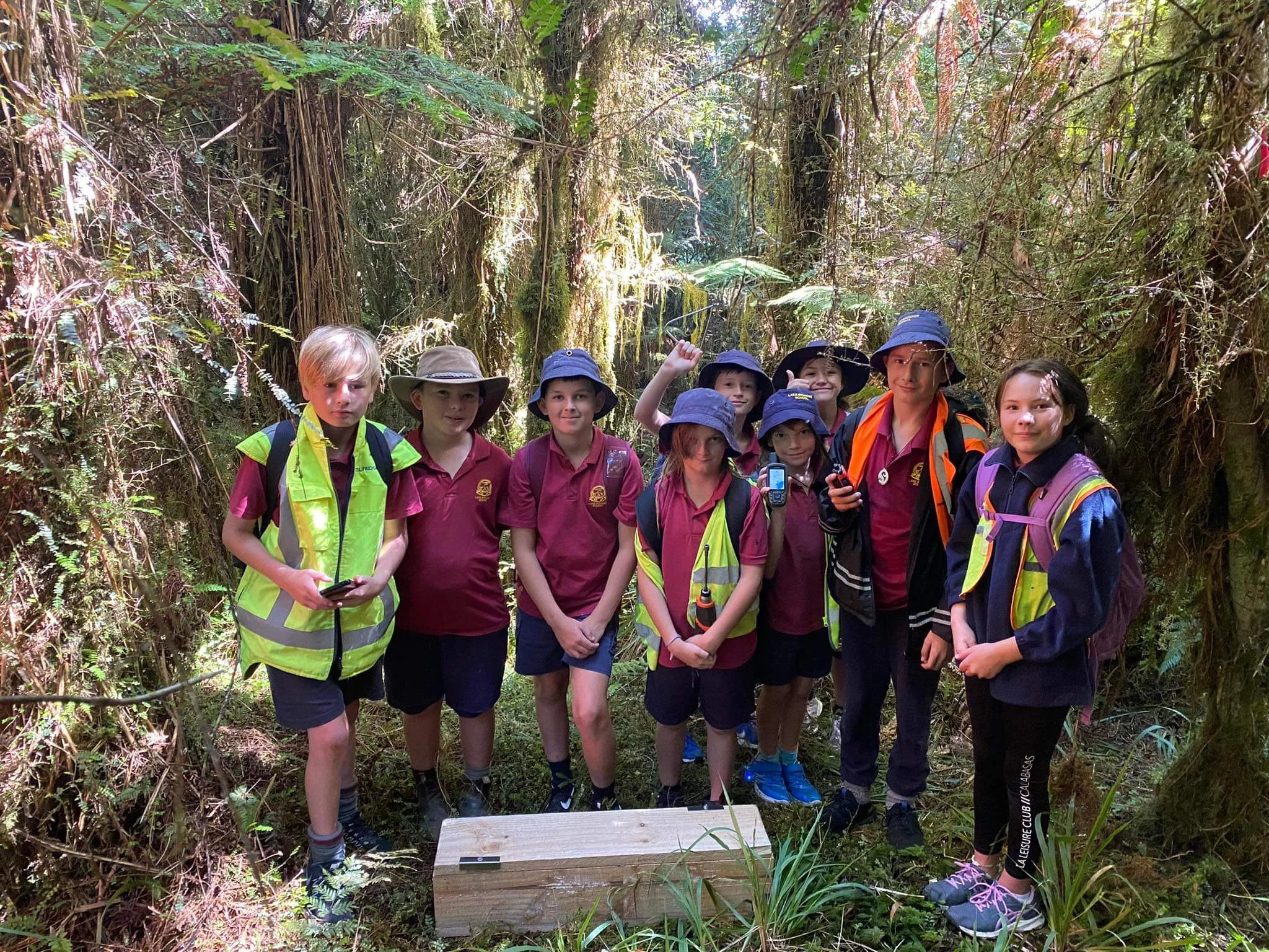Group of children on a nature walk in a dense forest, some wearing hats and backpacks, with one holding a GPS device and another with a radio, standing in front of lush greenery and moss-covered trees.