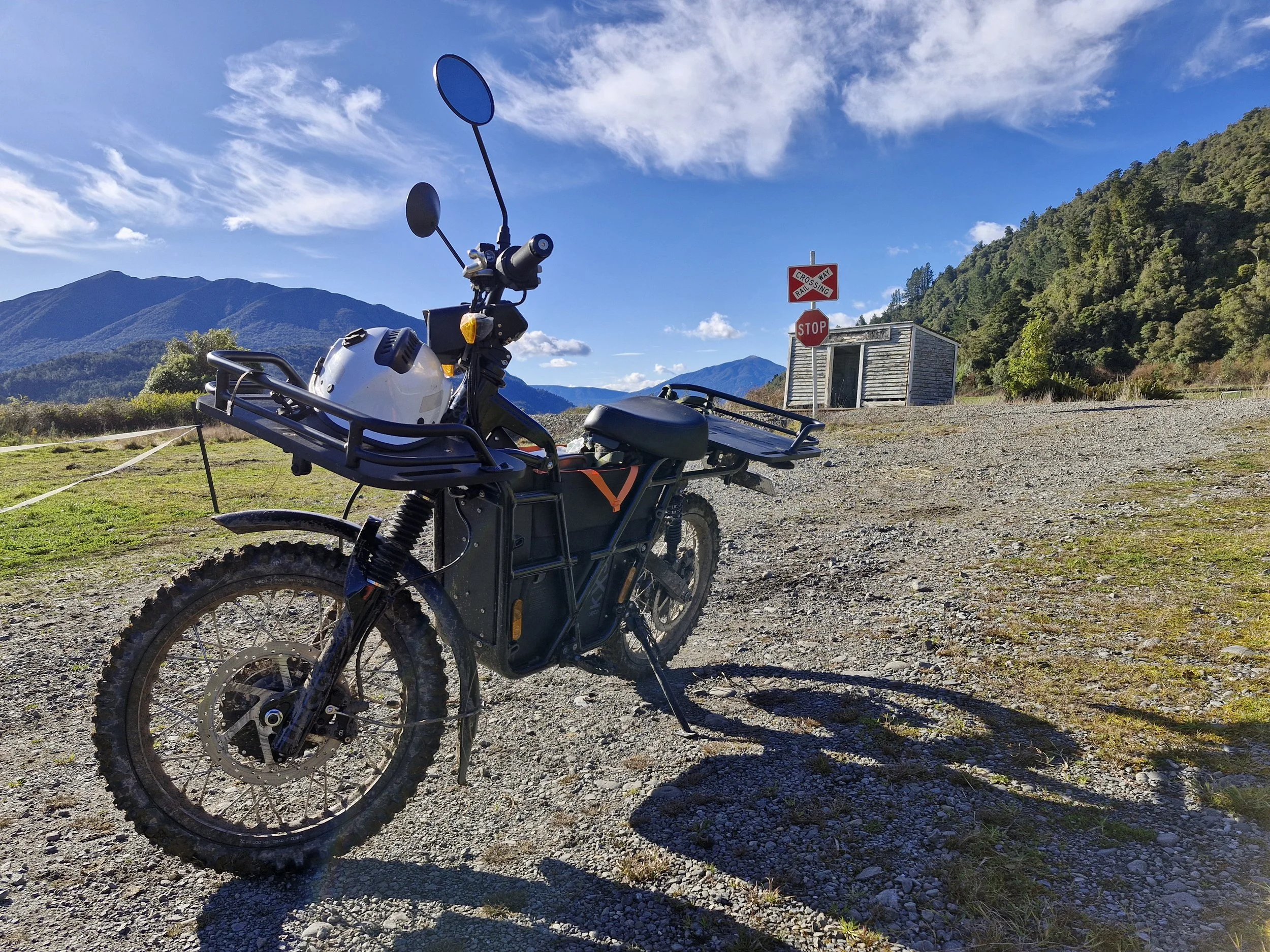 Electric bike parked on gravel road with mountains in the background, blue sky, and a small building with stop and crossing signs nearby.