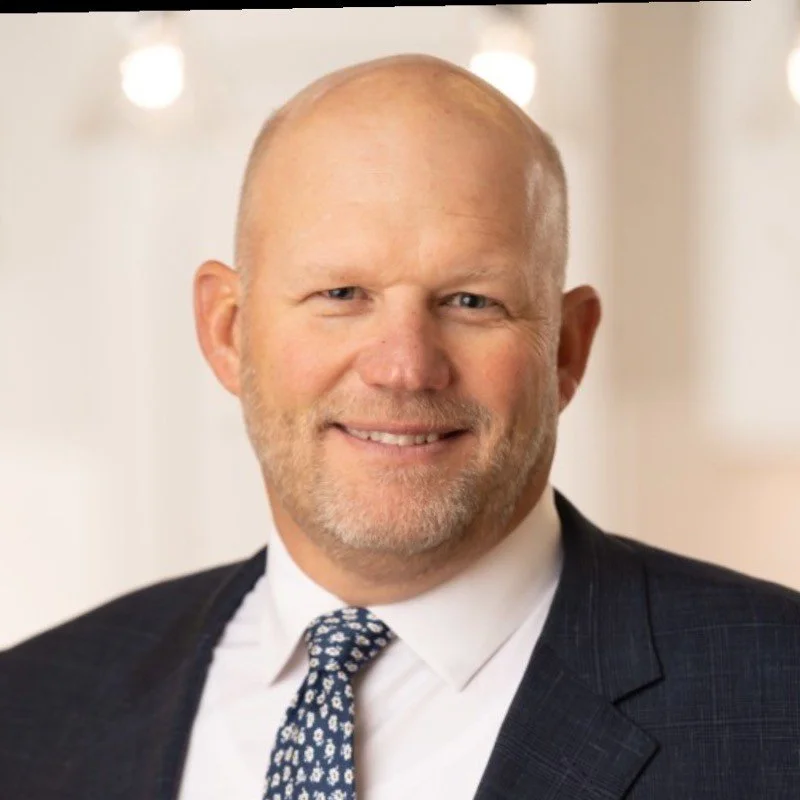 Portrait of a middle-aged man with a smile, wearing a navy suit, white shirt, and patterned tie, with a blurred background of lights.