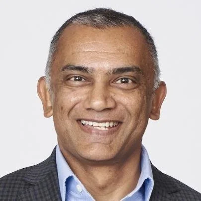A smiling man with short gray hair, wearing a dark blazer over a light blue shirt, against a plain light background.
