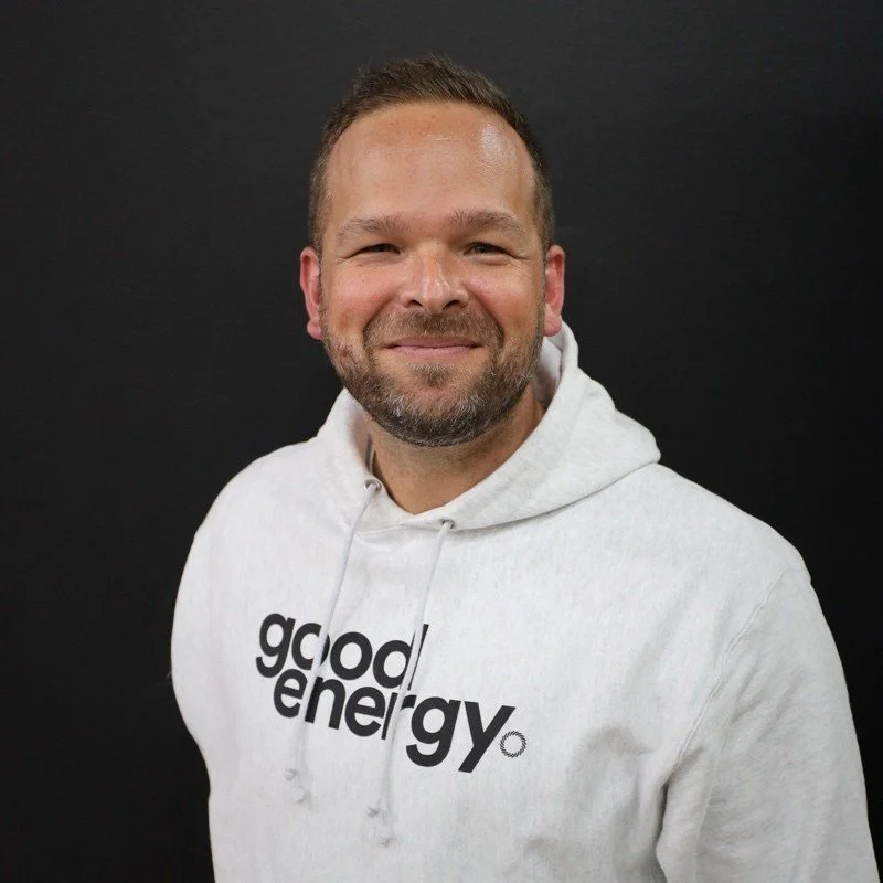 A smiling man wearing a white hoodie with 'good energy' text in black, standing against a dark background.