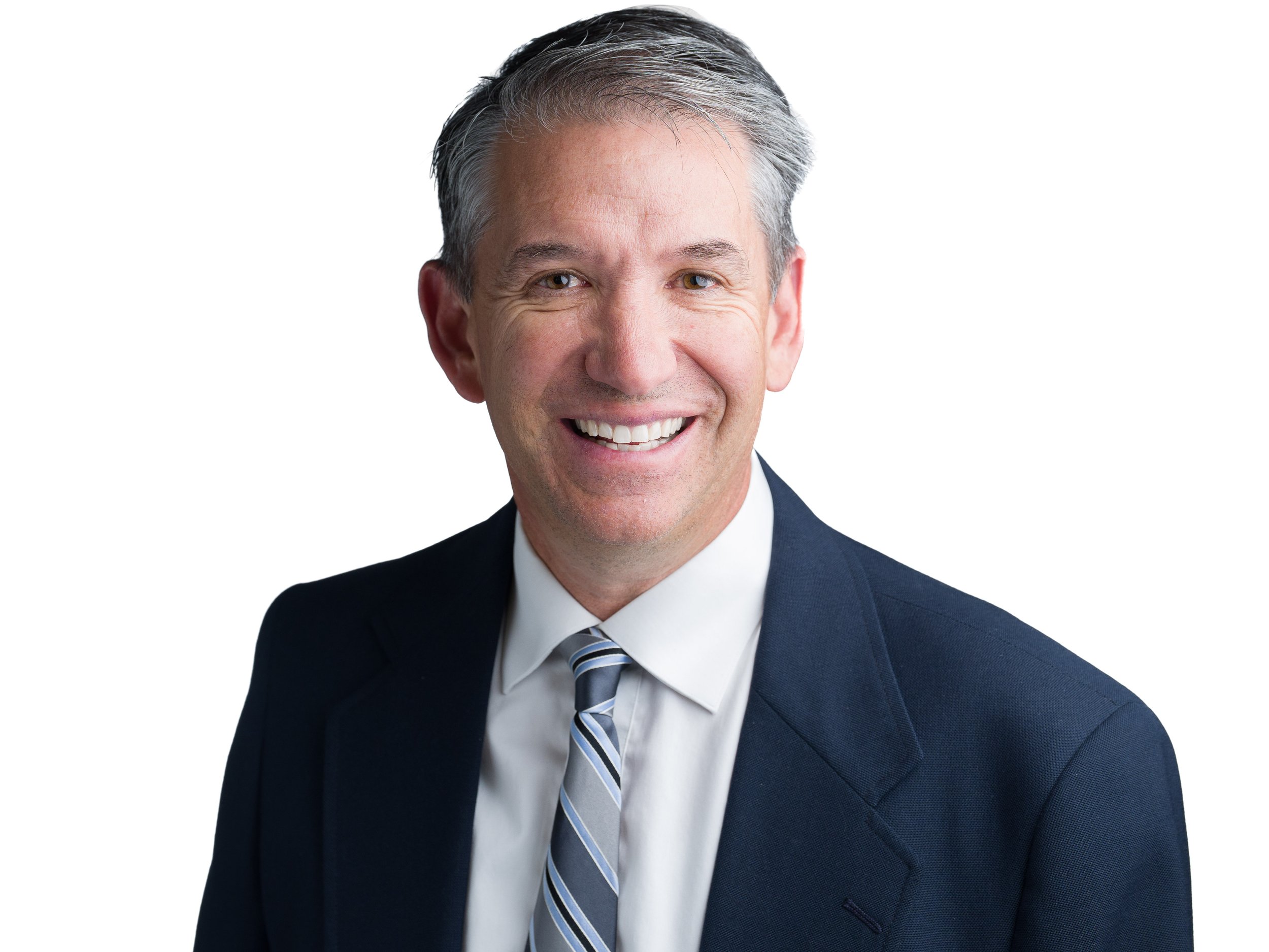A middle-aged man with gray hair, wearing a dark suit, white shirt, and tie, smiling in front of a light-colored background.