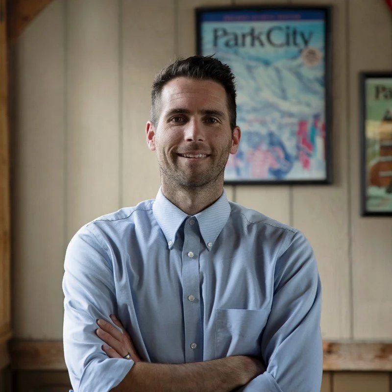 A man standing indoors with arms crossed, smiling, in front of a wall with framed posters, including one that says "Park City".