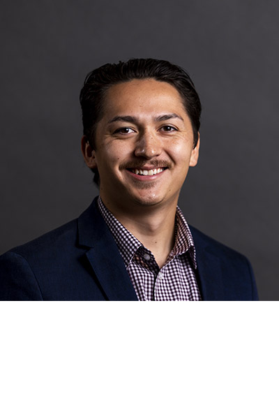 A smiling man in a dark blazer and checkered shirt against a gray background.