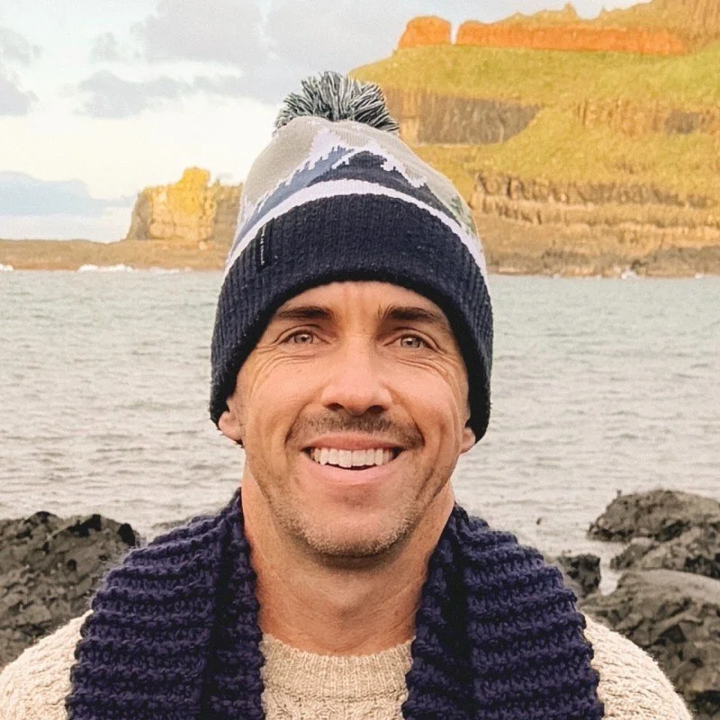 Smiling man outdoors near the water, wearing a winter hat and scarf, with cliffs in the background.