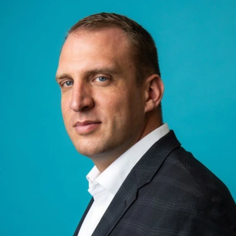 Portrait of a man in a dark suit jacket with a white shirt against a teal background.