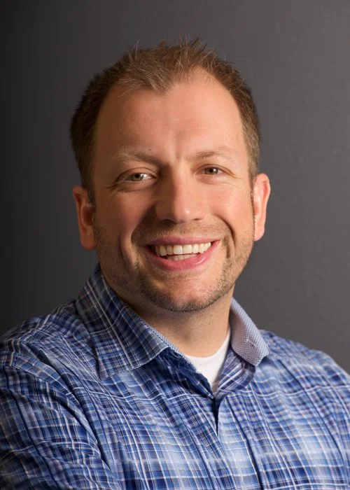 A smiling man in a blue plaid shirt with short, light brown hair posing against a gray background.