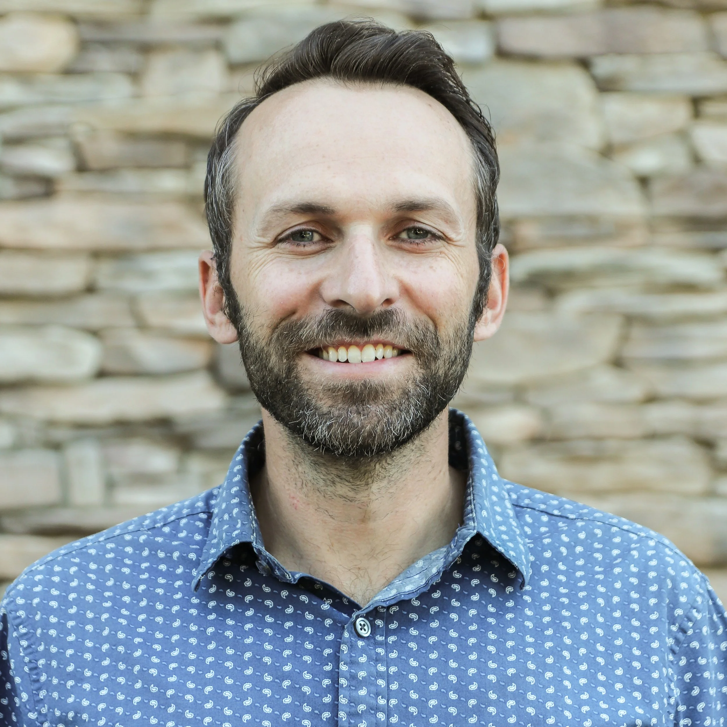 Close-up of a smiling man with a beard and short brown hair, outdoors with blurred green trees in the background.