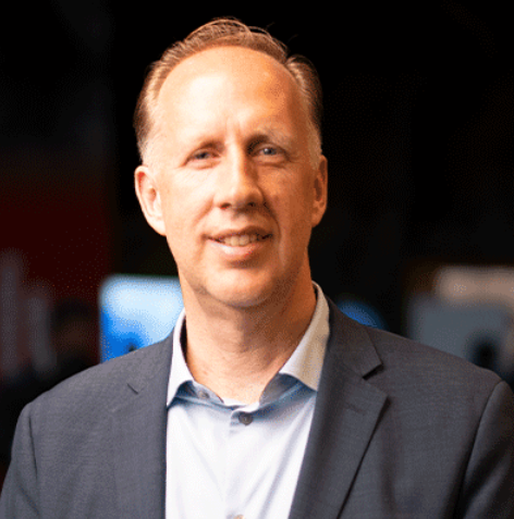 A professional man in a suit smiling in a dimly lit indoor setting.