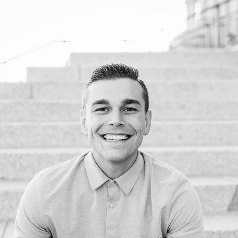 Smiling young man with short neatly styled hair, wearing a light-colored collared shirt, sitting on outdoor stairs.