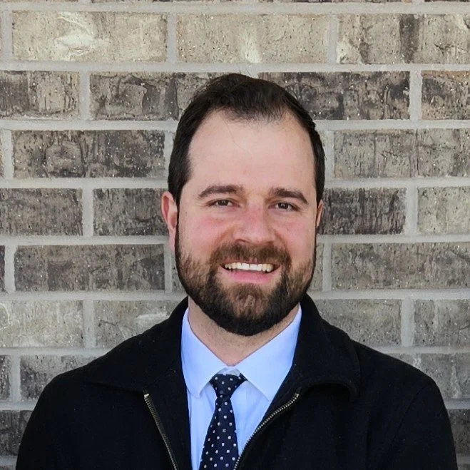 A man with a beard and short brown hair smiling, wearing a white dress shirt, blue patterned tie, and black jacket, standing in front of a brick wall.