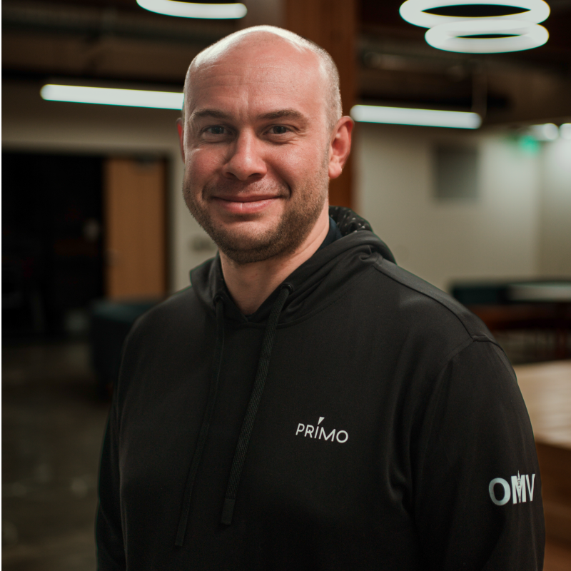 A smiling man with a shaved head and beard in a black hoodie with 'PRIMO' and 'OMV' logos, standing indoors with modern lighting fixtures.
