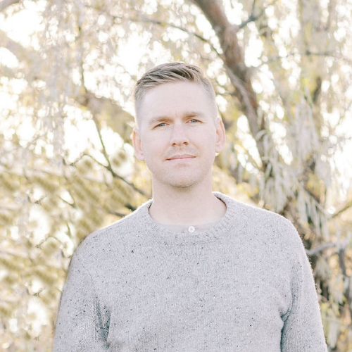 A young man with short hair smiling and standing outdoors with trees in the background, wearing a maroon button-up shirt with arms crossed.