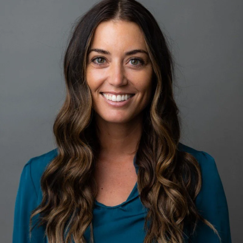 A smiling woman with long wavy brown hair, wearing a teal blouse, standing against a gray background.