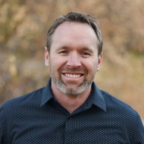 A smiling man with short brown hair and a beard, wearing a navy blue, dotted button-up shirt, outdoors in a fall setting.