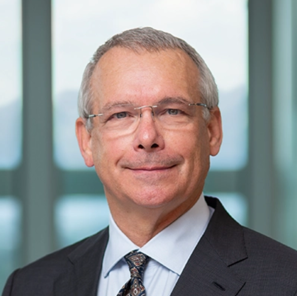 A middle-aged man with gray hair and glasses, dressed in a suit and tie, standing in front of a large window with a blurred cityscape background.
